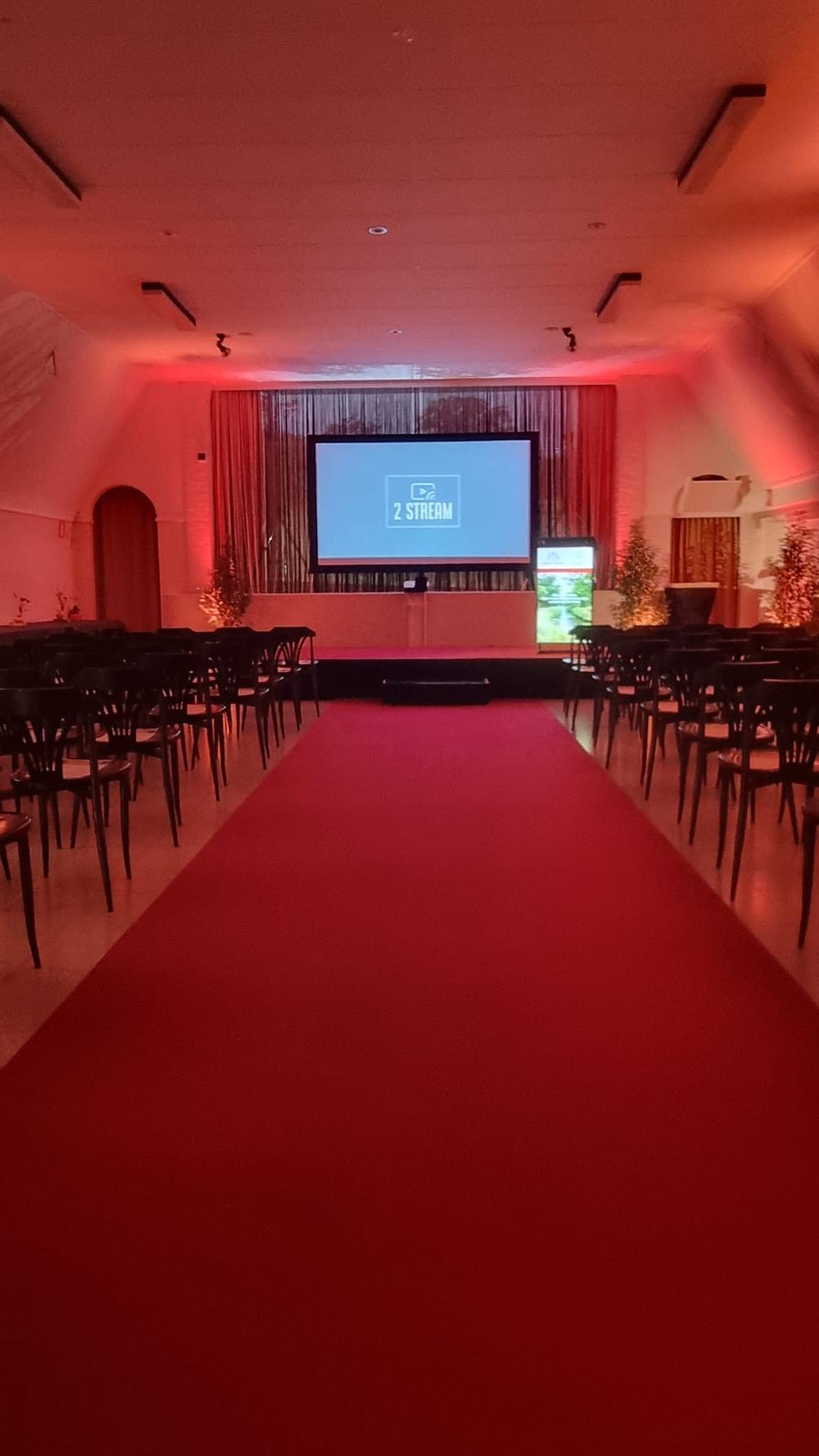 Hall with rows of chairs and a red carpet leading to a stage screen showing the 2 Stream logo