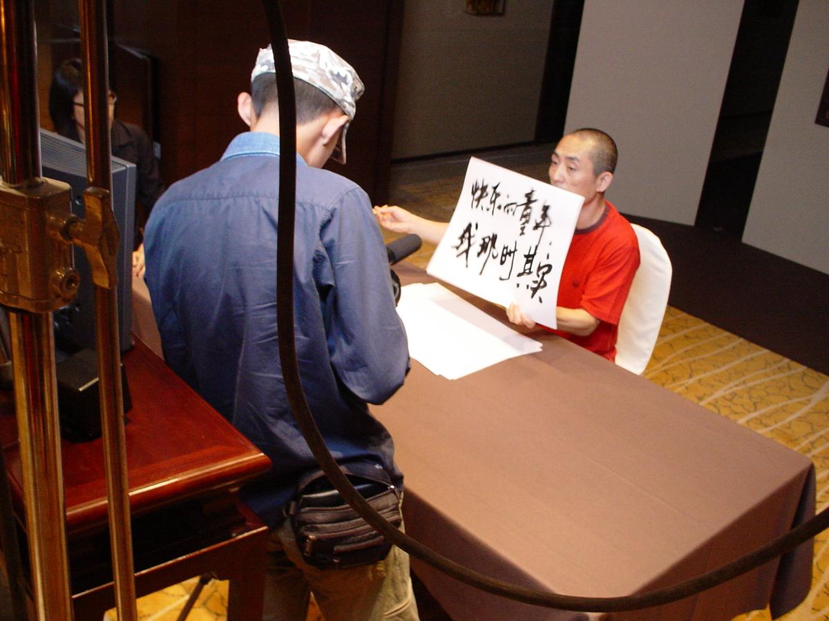 Camera operator filming a man in a red shirt holding up two lines of Chinese calligraphy