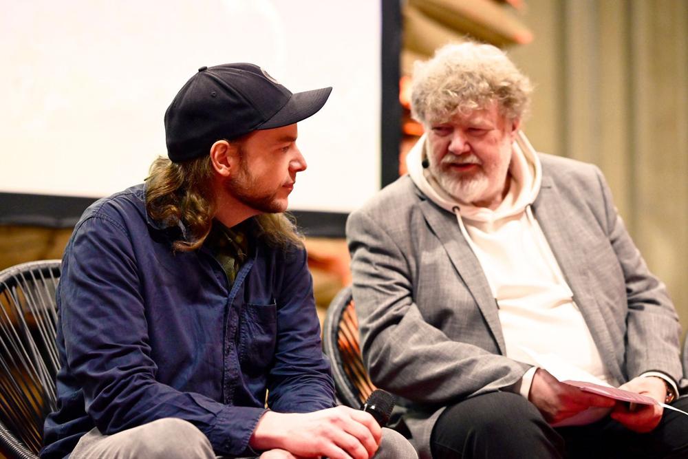 Two men seated on stage in conversation, one in a cap holding a microphone, the other in a grey blazer