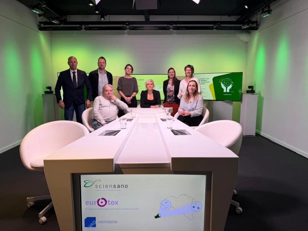 Group posing around a white desk in a green-lit studio, with Sciensano, Eurotox and VAD logos on the desk screen