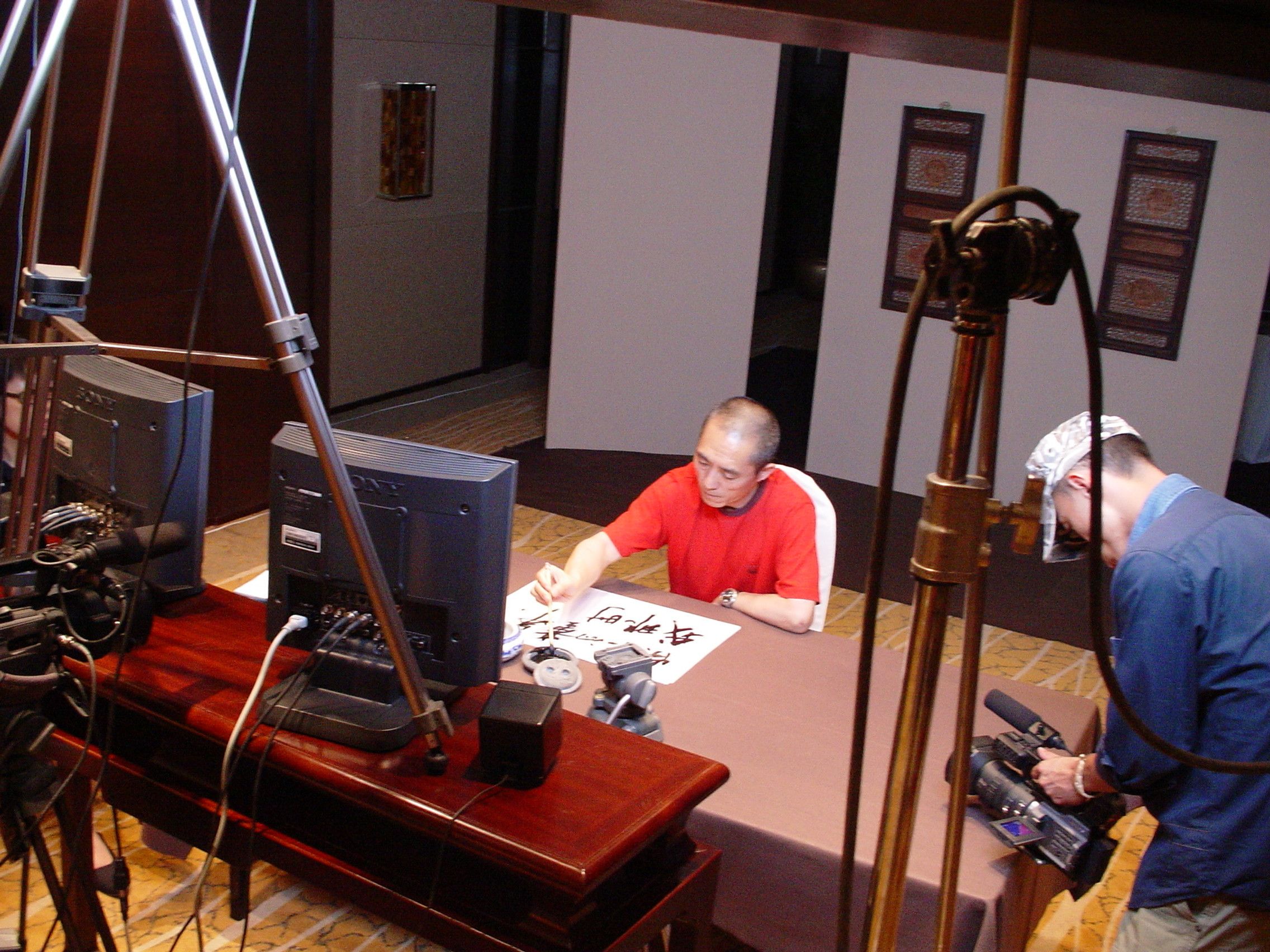 Man in red writing calligraphy at a table on a hotel set, surrounded by monitors, light stands and a camera