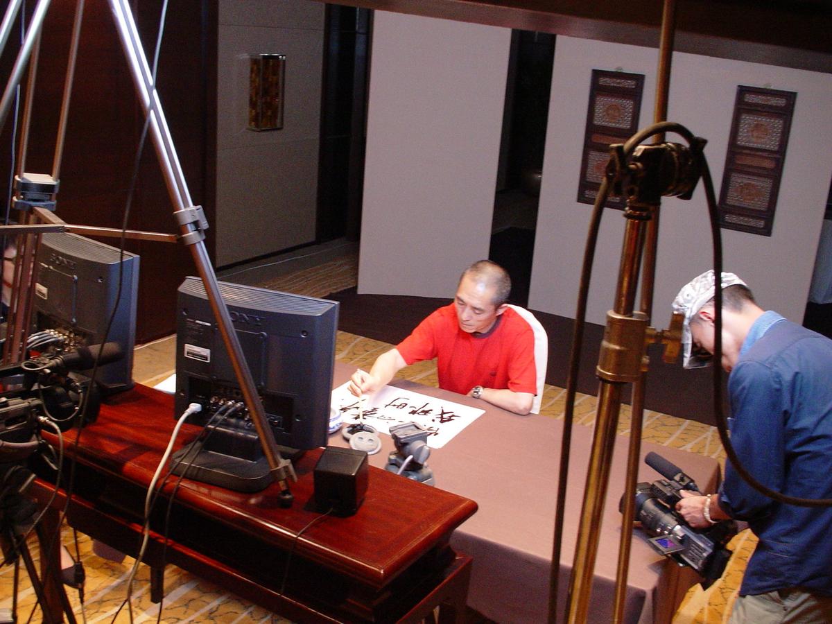 Man in red writing calligraphy at a table on a hotel set, surrounded by monitors, light stands and a camera