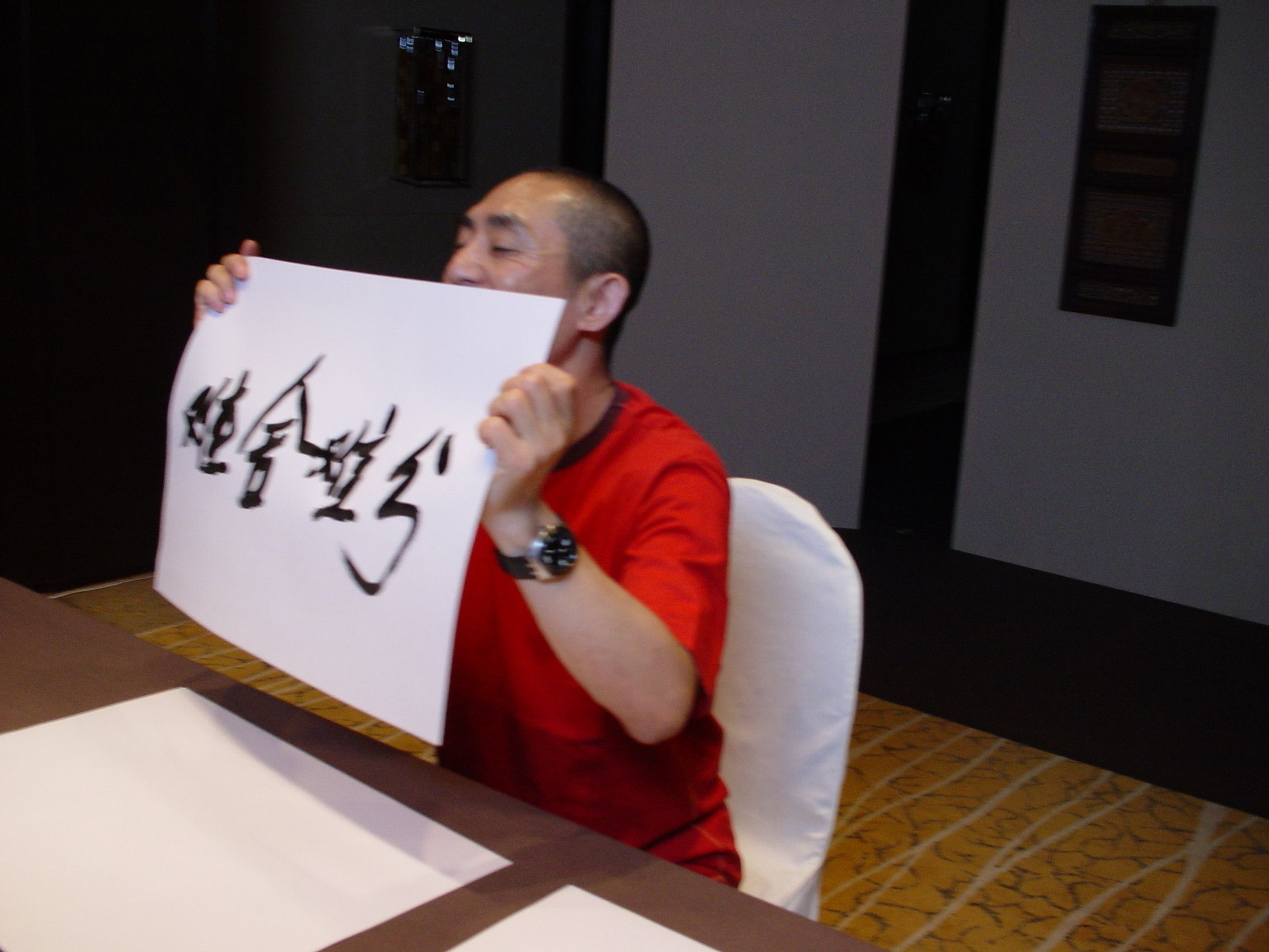 Man in a red T-shirt seated at a table, smiling and holding up a sheet of Chinese calligraphy