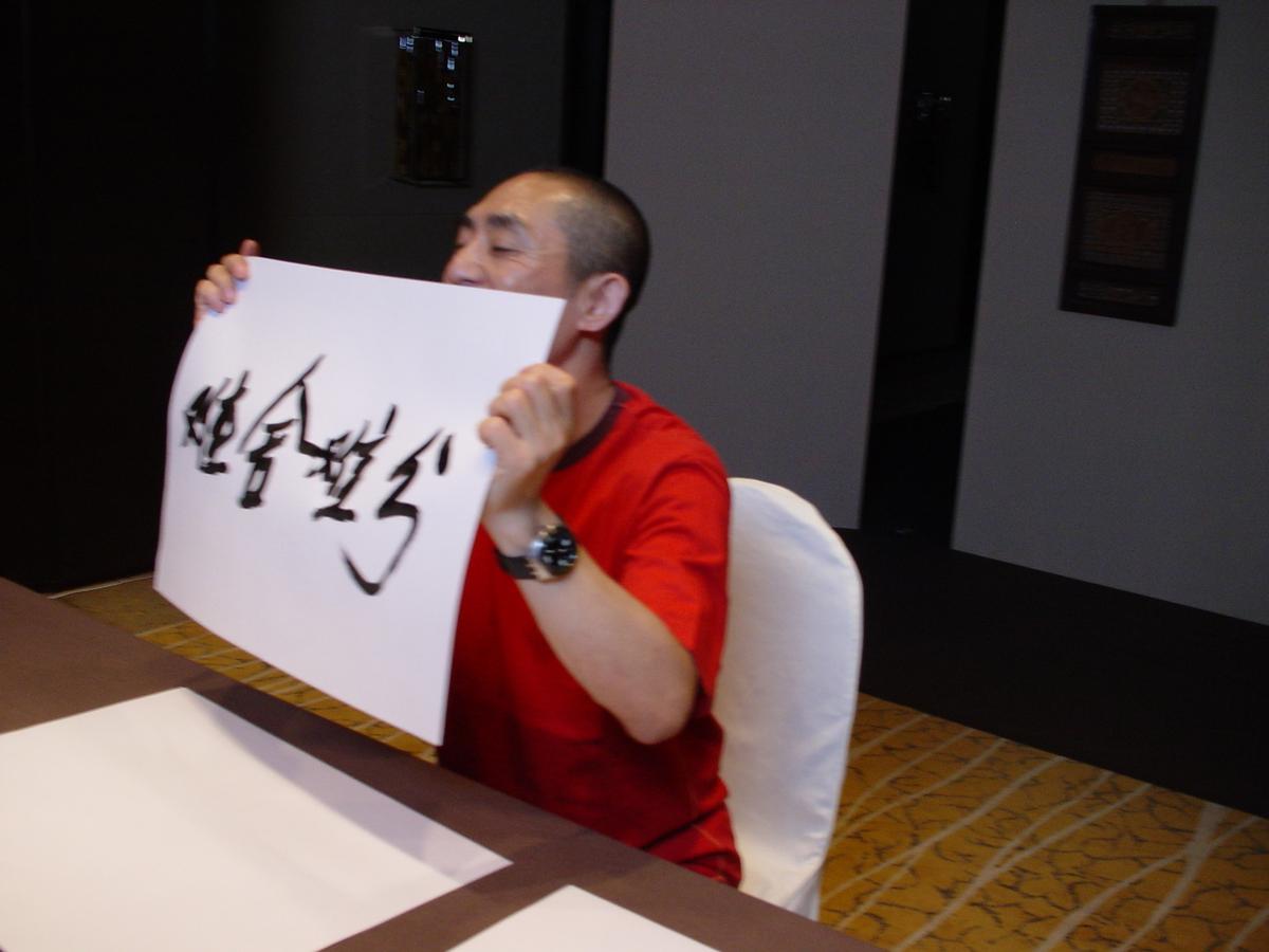 Man in a red T-shirt seated at a table, smiling and holding up a sheet of Chinese calligraphy