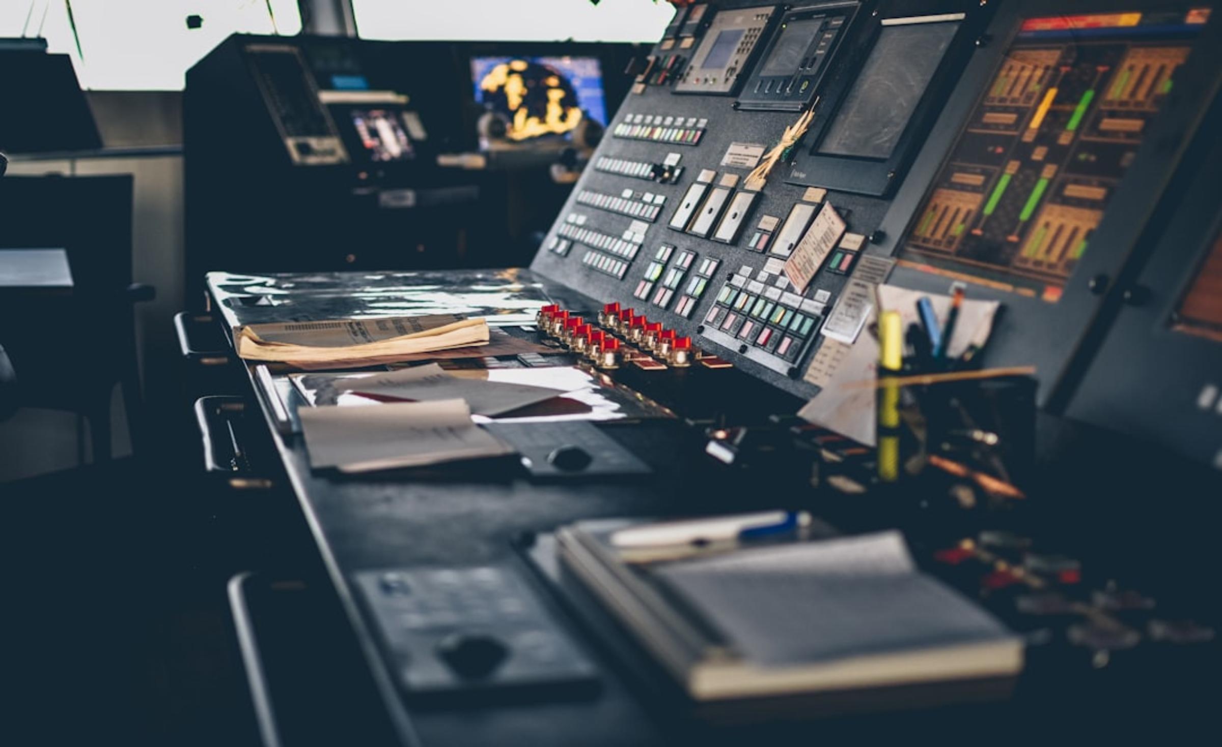 Dimly lit control console with rows of buttons, red switches and level meters, papers and a notebook on the desk