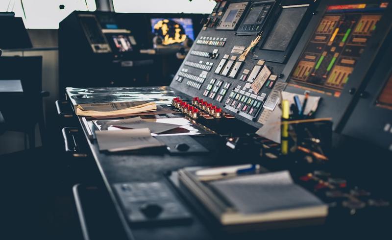 Dimly lit control console with rows of buttons, red switches and level meters, papers and a notebook on the desk