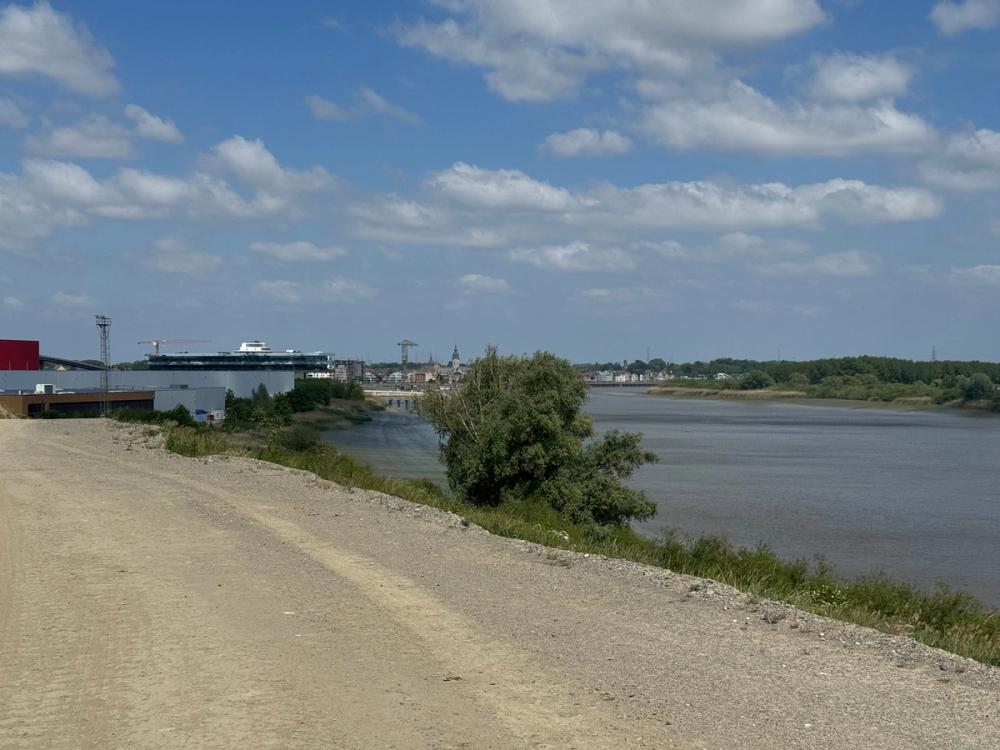 Gravel dyke path along a wide river, with warehouses, cranes and a church spire in the distance