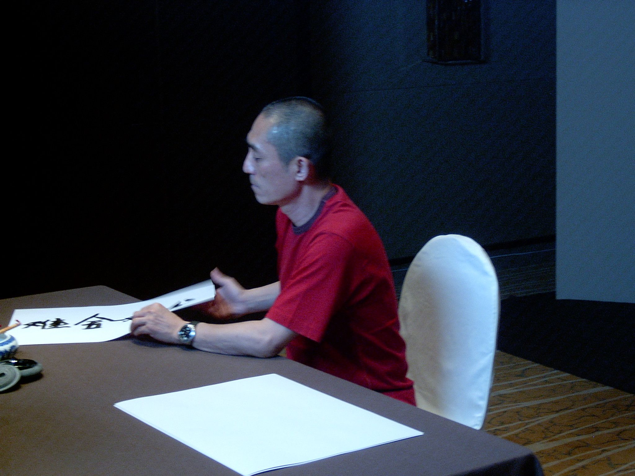 Man in a red T-shirt writing Chinese calligraphy with a brush at a table in a darkened room