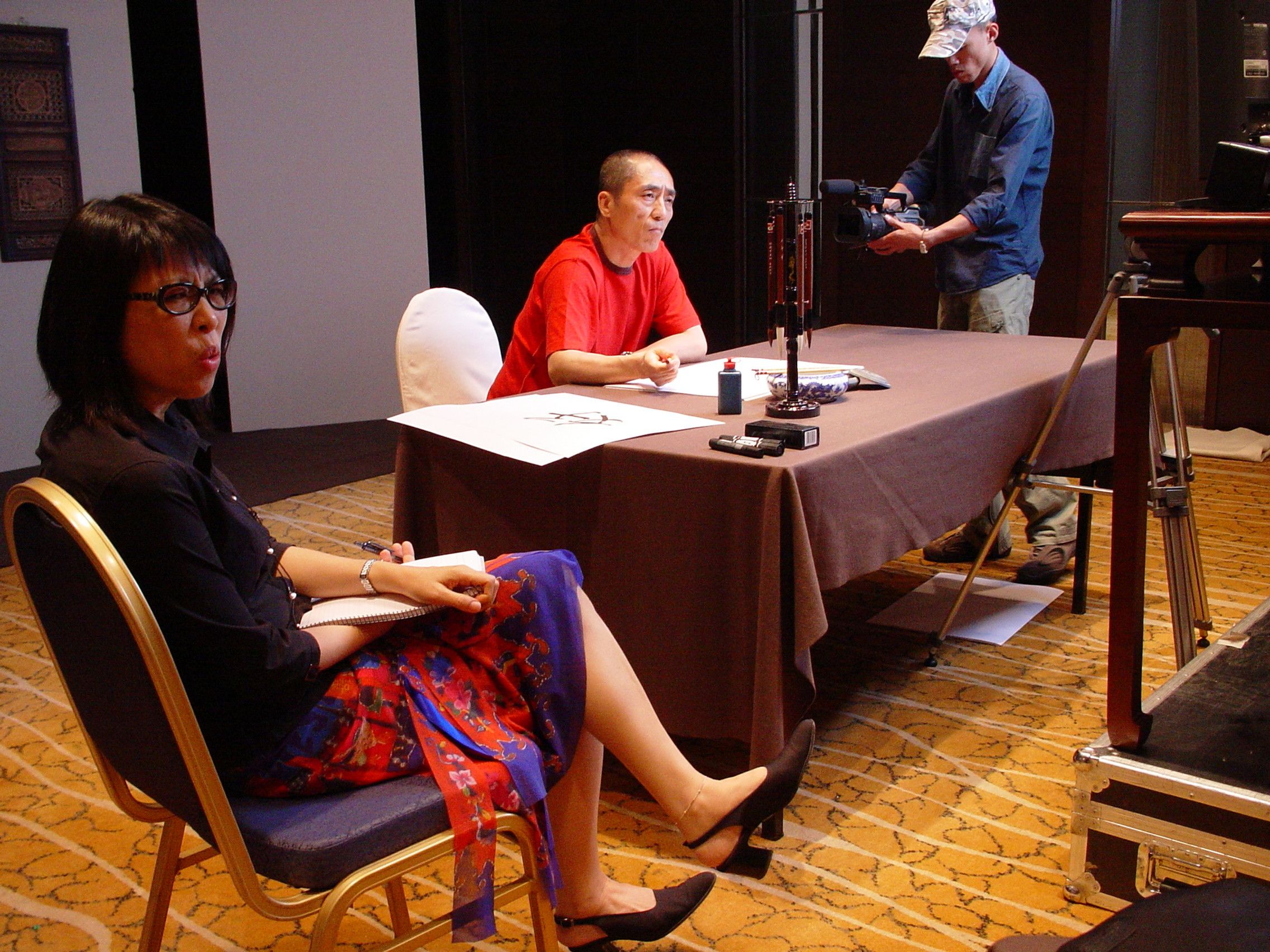 Woman taking notes in the foreground as a man in red sits at a calligraphy table and a camera operator films