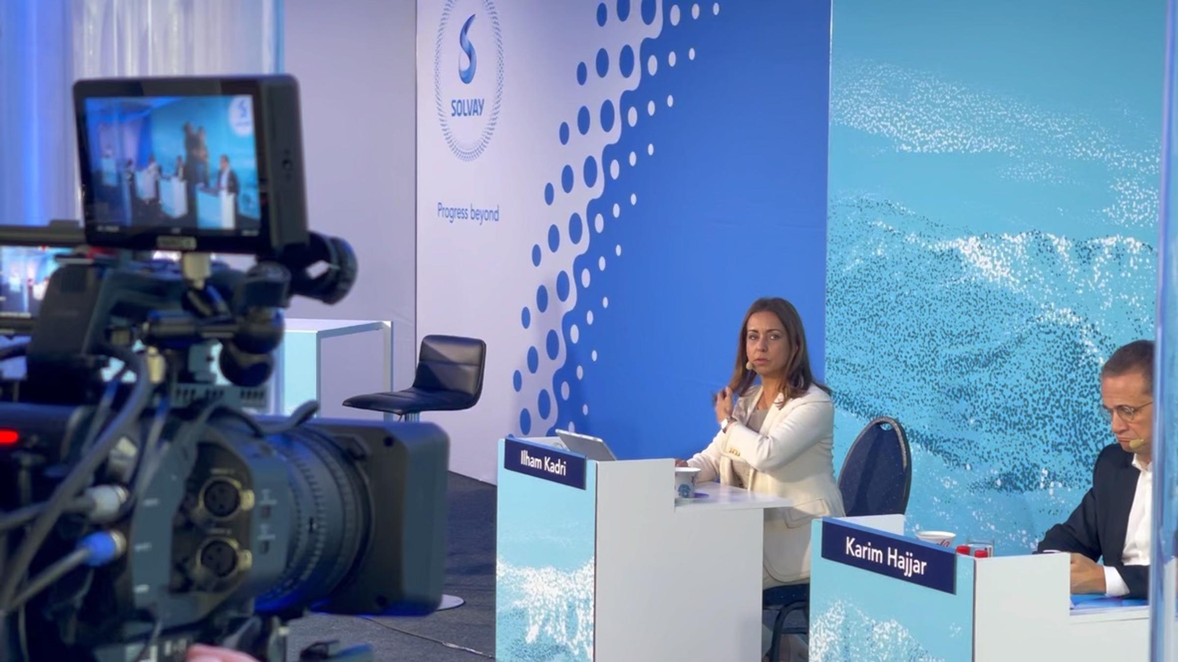 A broadcast camera trained on a speaker at a branded corporate set during a live stream