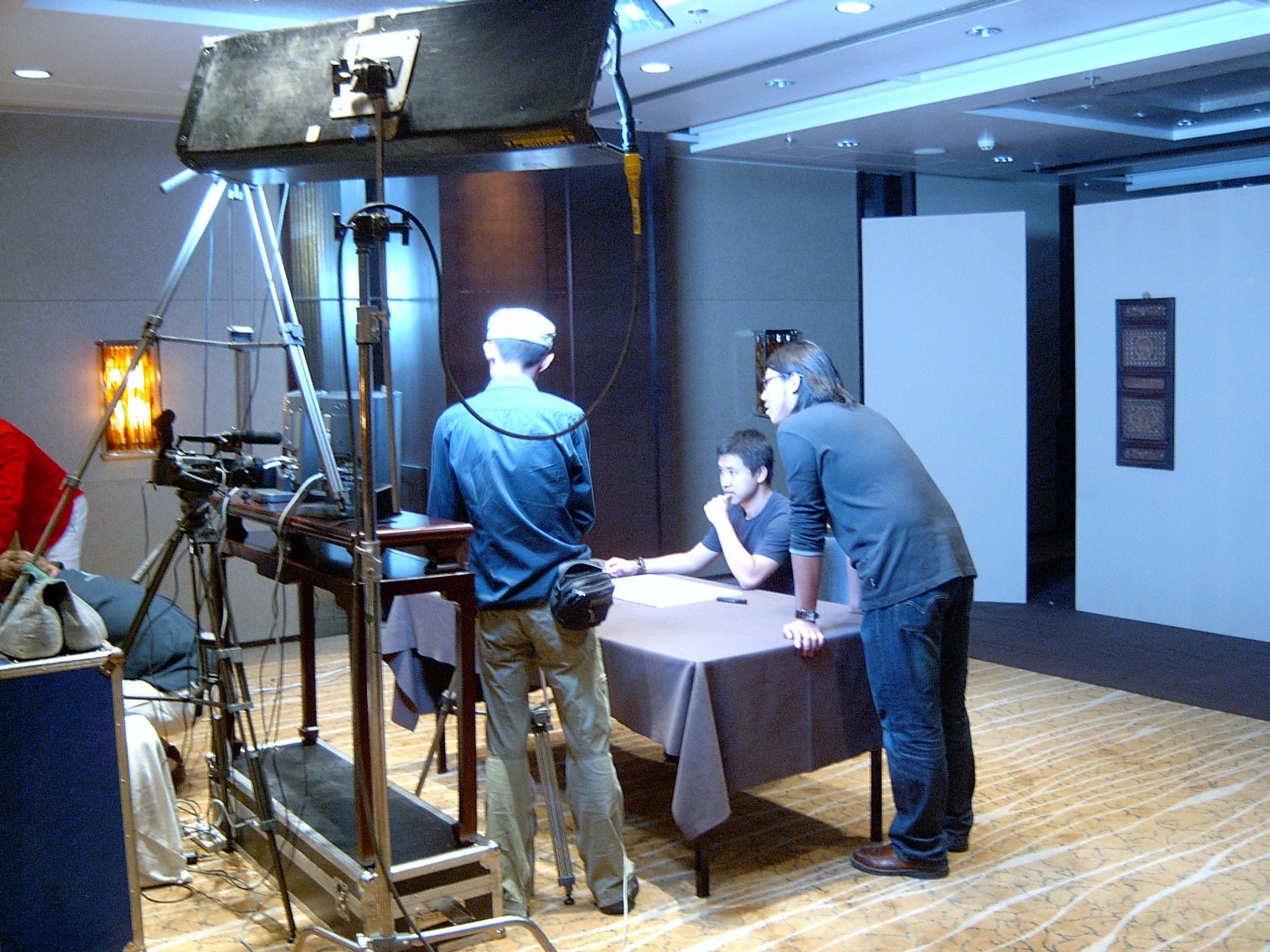 Crew rigging a camera and overhead light in a hotel function room while a man waits at a table on set