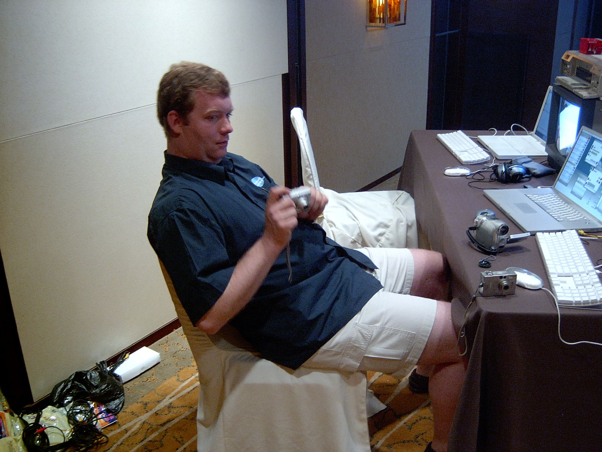 Crew member leaning back in a chair holding a compact camera beside a table of laptops and camcorders