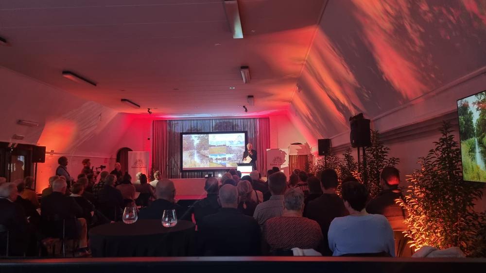 Audience watching a speaker at a lectern beside a screen showing a landscape, in a hall lit in red