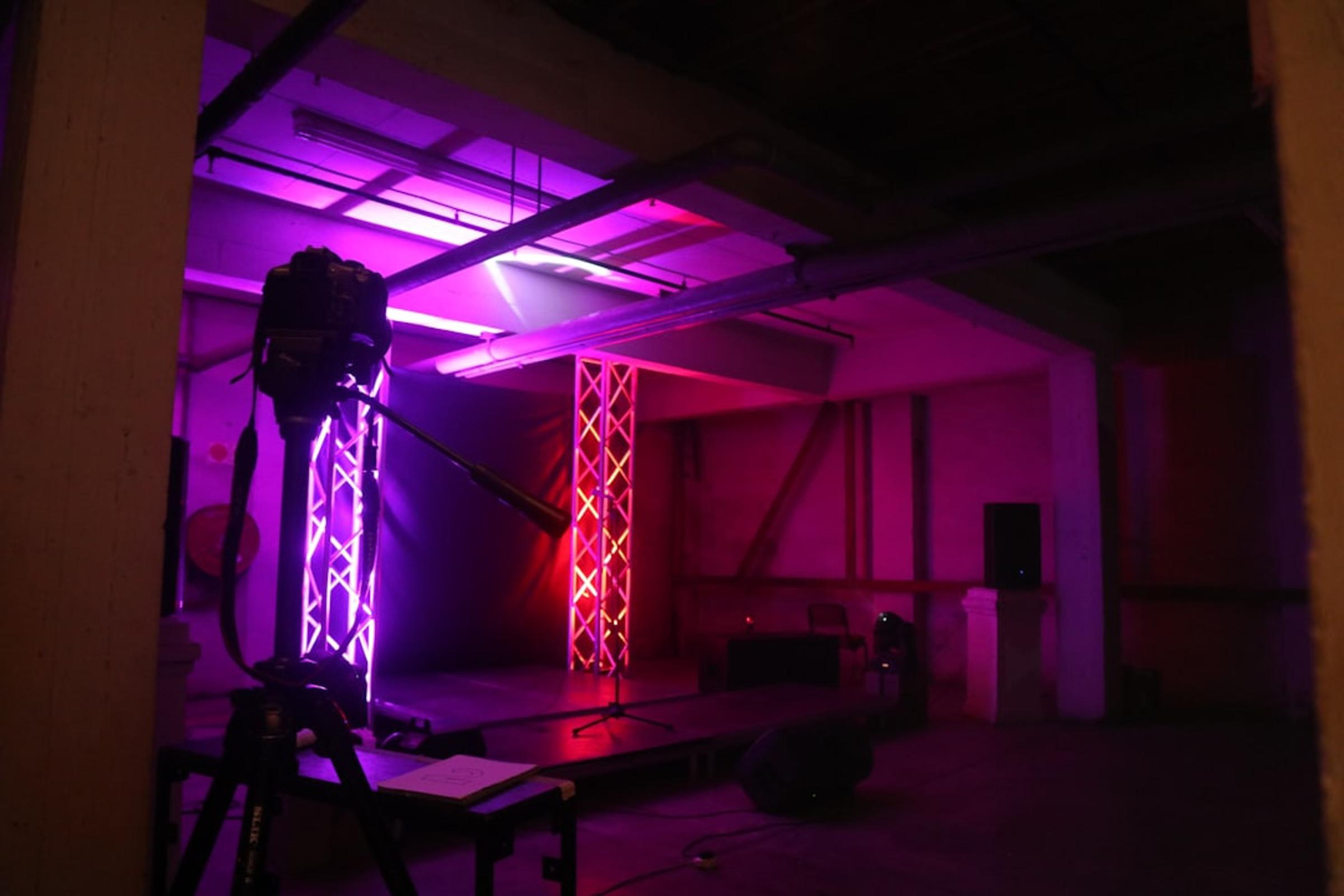Camera on a tripod facing a small stage lit in purple and pink in an empty industrial space