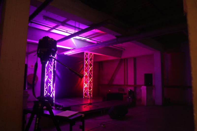 Camera on a tripod facing a small stage lit in purple and pink in an empty industrial space