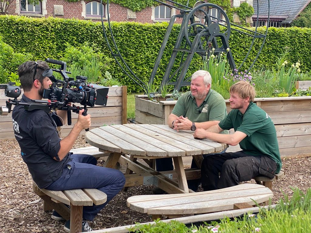 A camera operator filming an interview with two people at an outdoor table on location