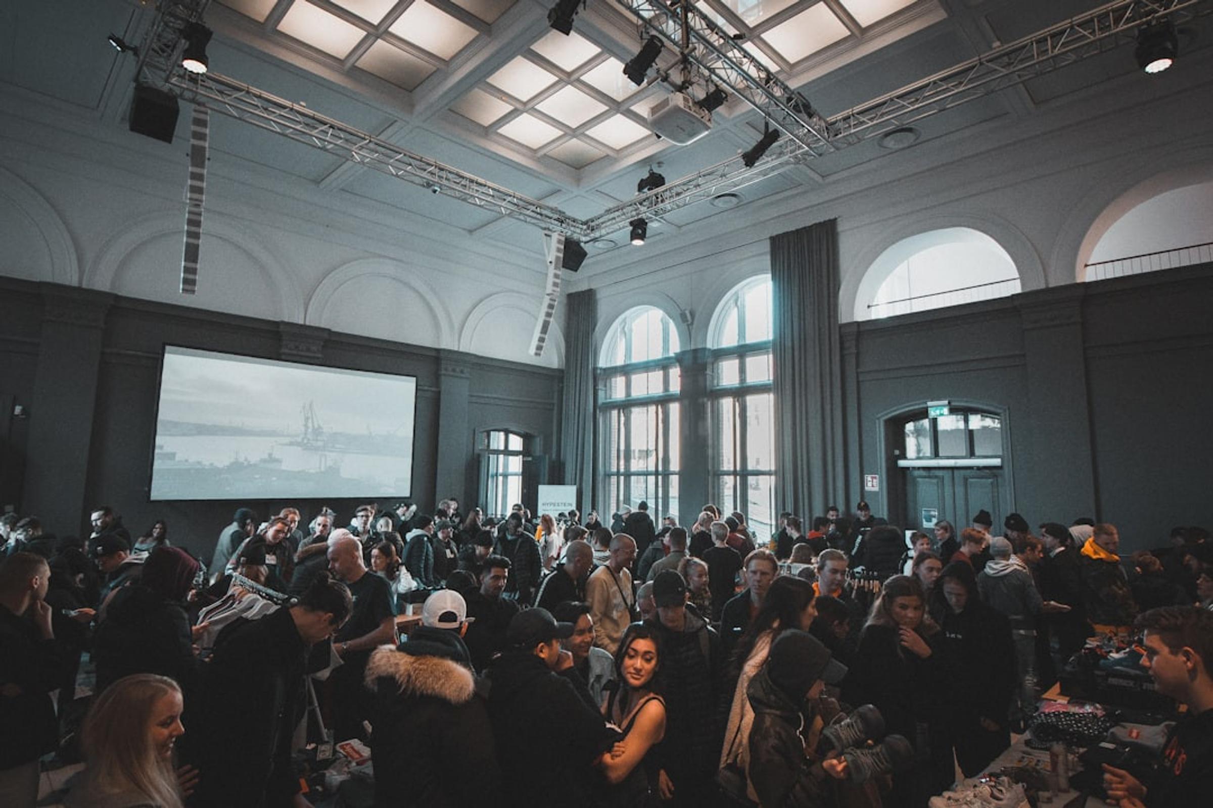 Crowded indoor event in a hall with arched windows, lighting trusses and a large projection screen