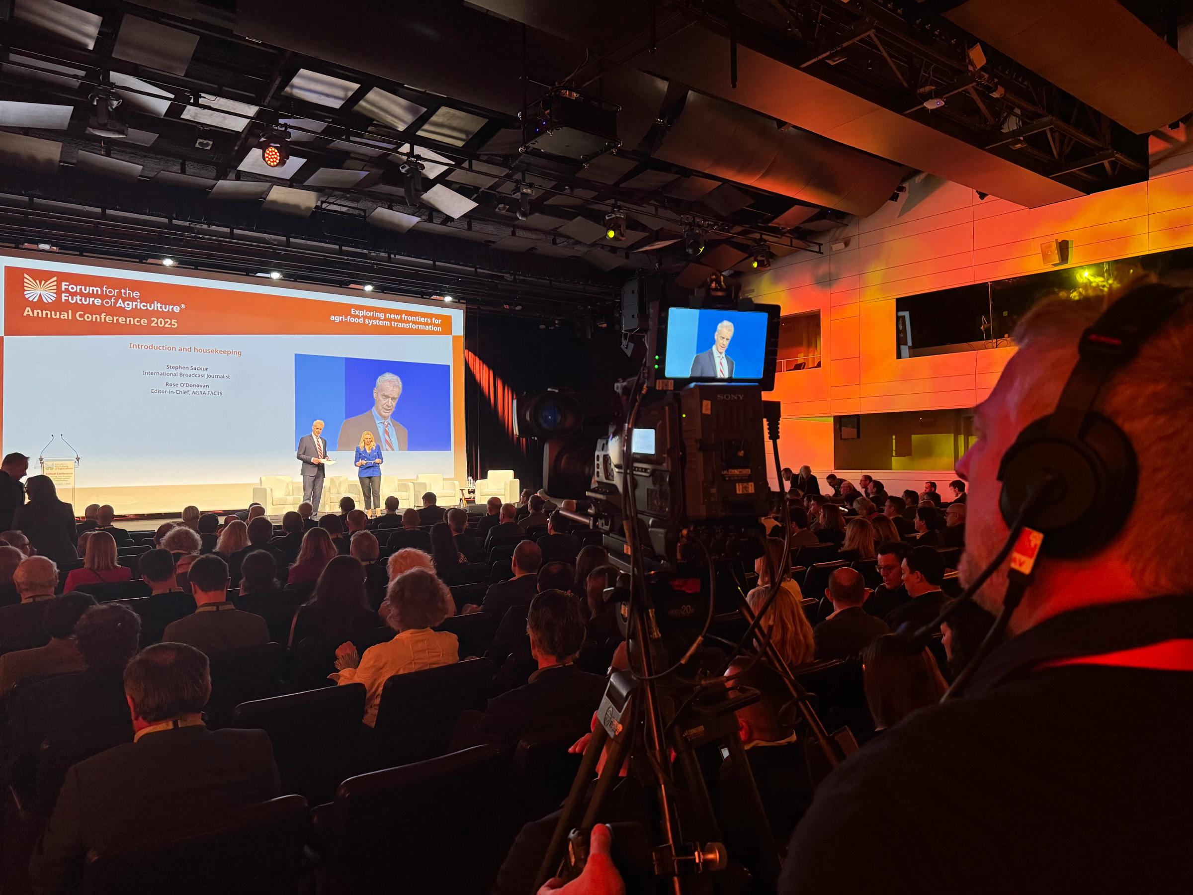 A camera operator filming a conference from the back of a full auditorium, the stage lit ahead