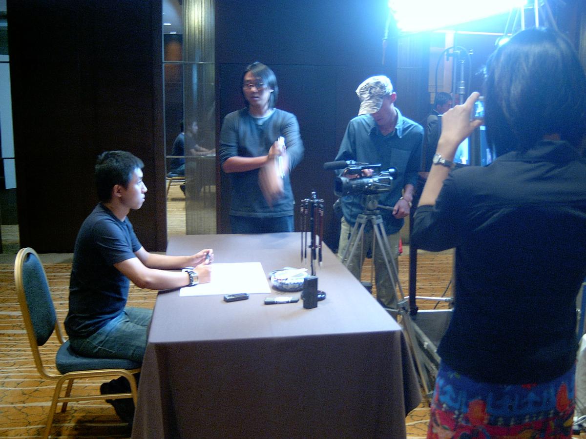 Young man seated at a calligraphy table while a camera operator and crew set up the shot