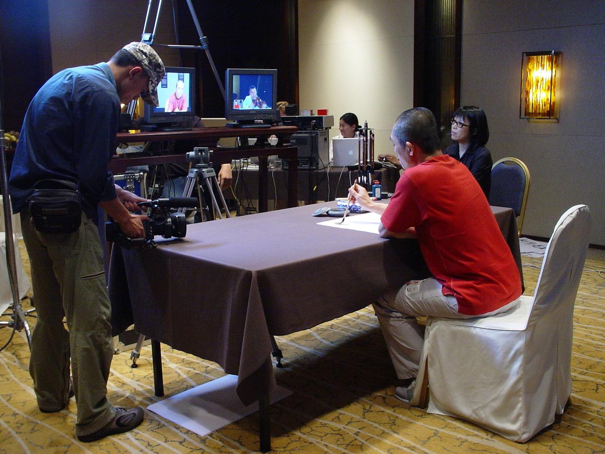 Camera operator filming a man in red writing calligraphy at a table, with preview monitors in the background