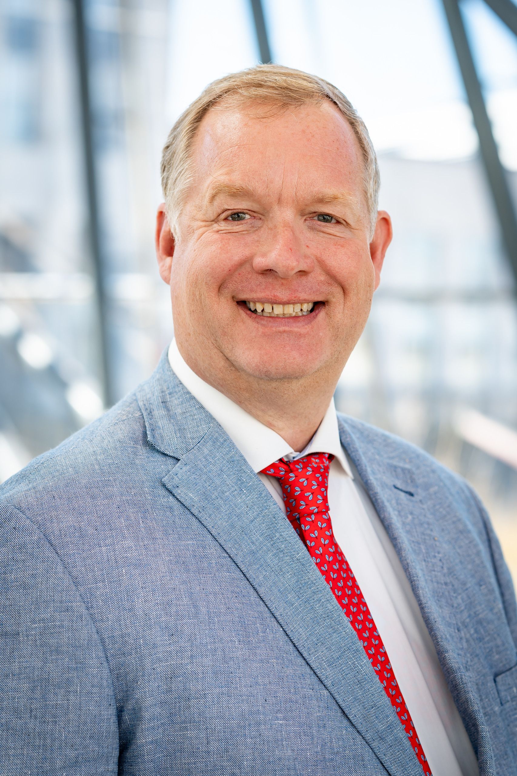 Portrait of Christophe Lenaerts smiling, in a light blue blazer and red patterned tie