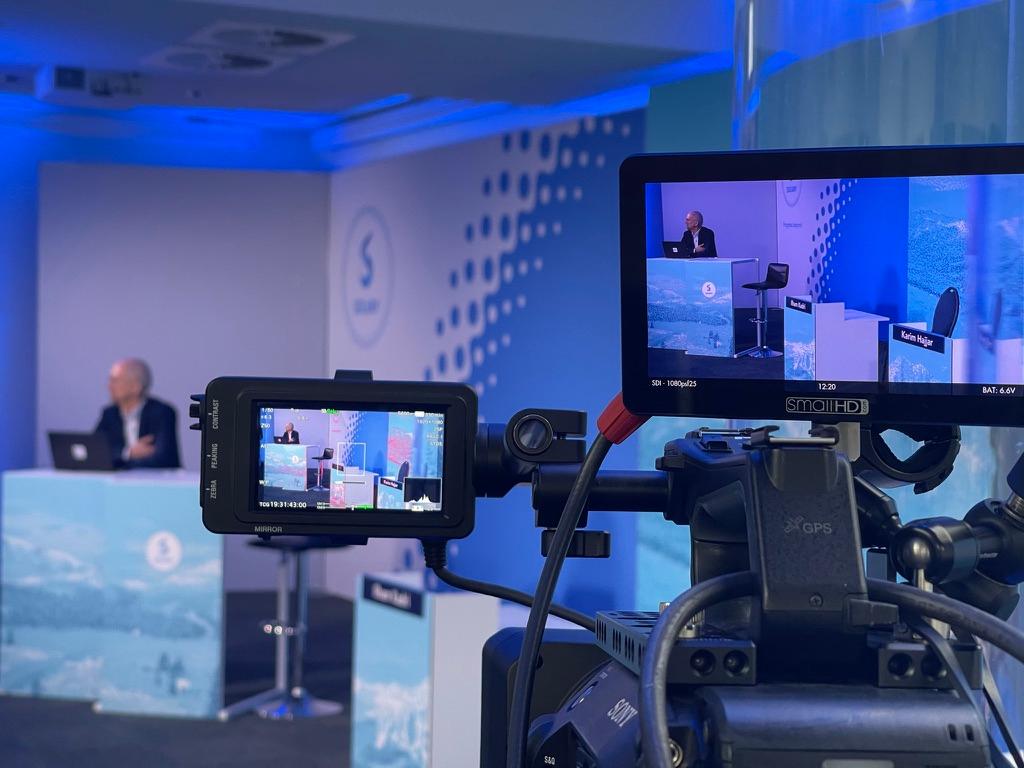 A broadcast camera and its monitor framing a speaker on a branded corporate set