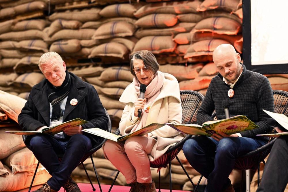 Three readers with microphones reading picture books aloud on stage in front of stacked jute sacks