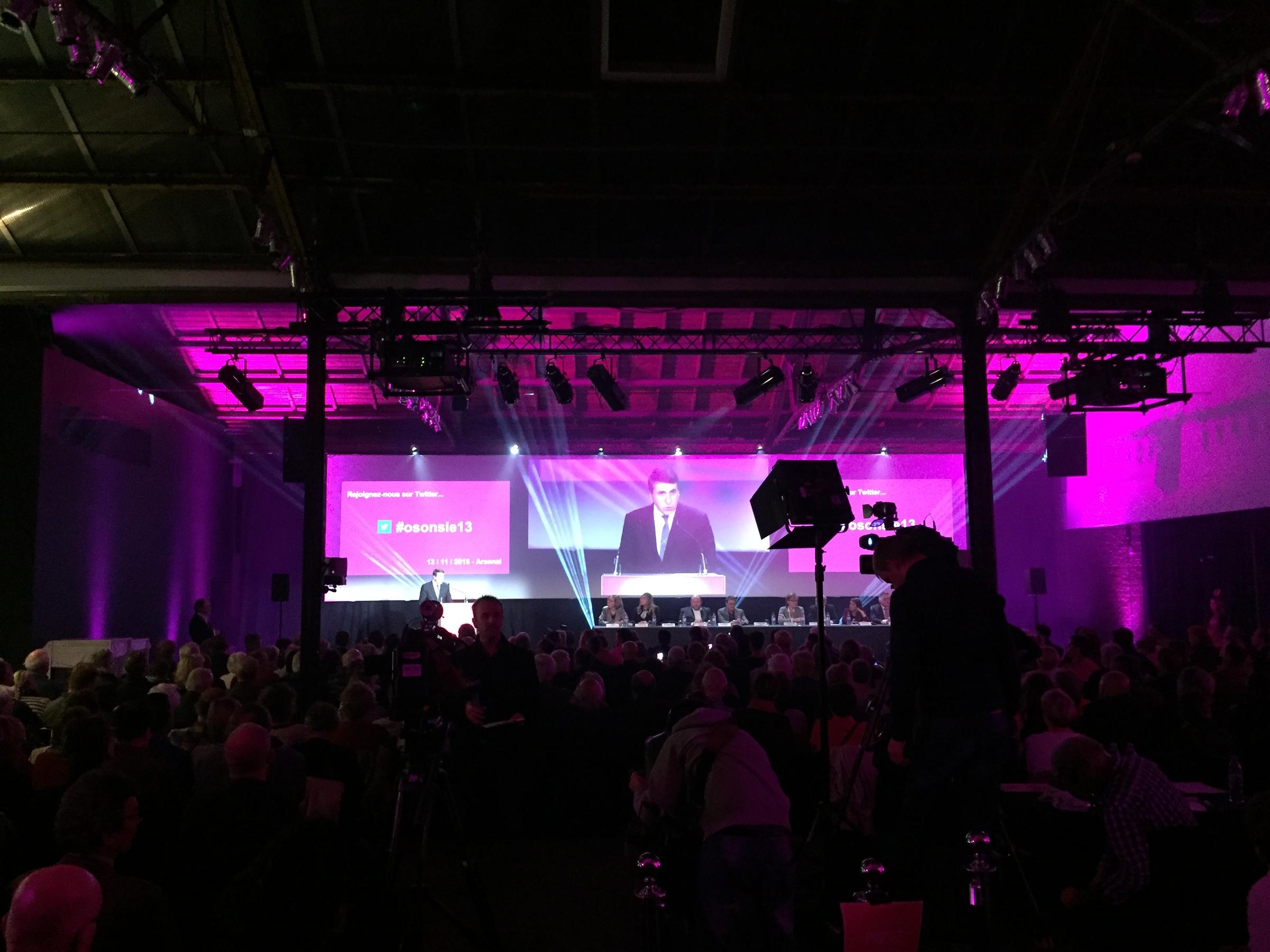 A conference hall filled with several hundred attendees watching a speaker on a large screen, with a 2 Stream camera filming the stage from the back of the room