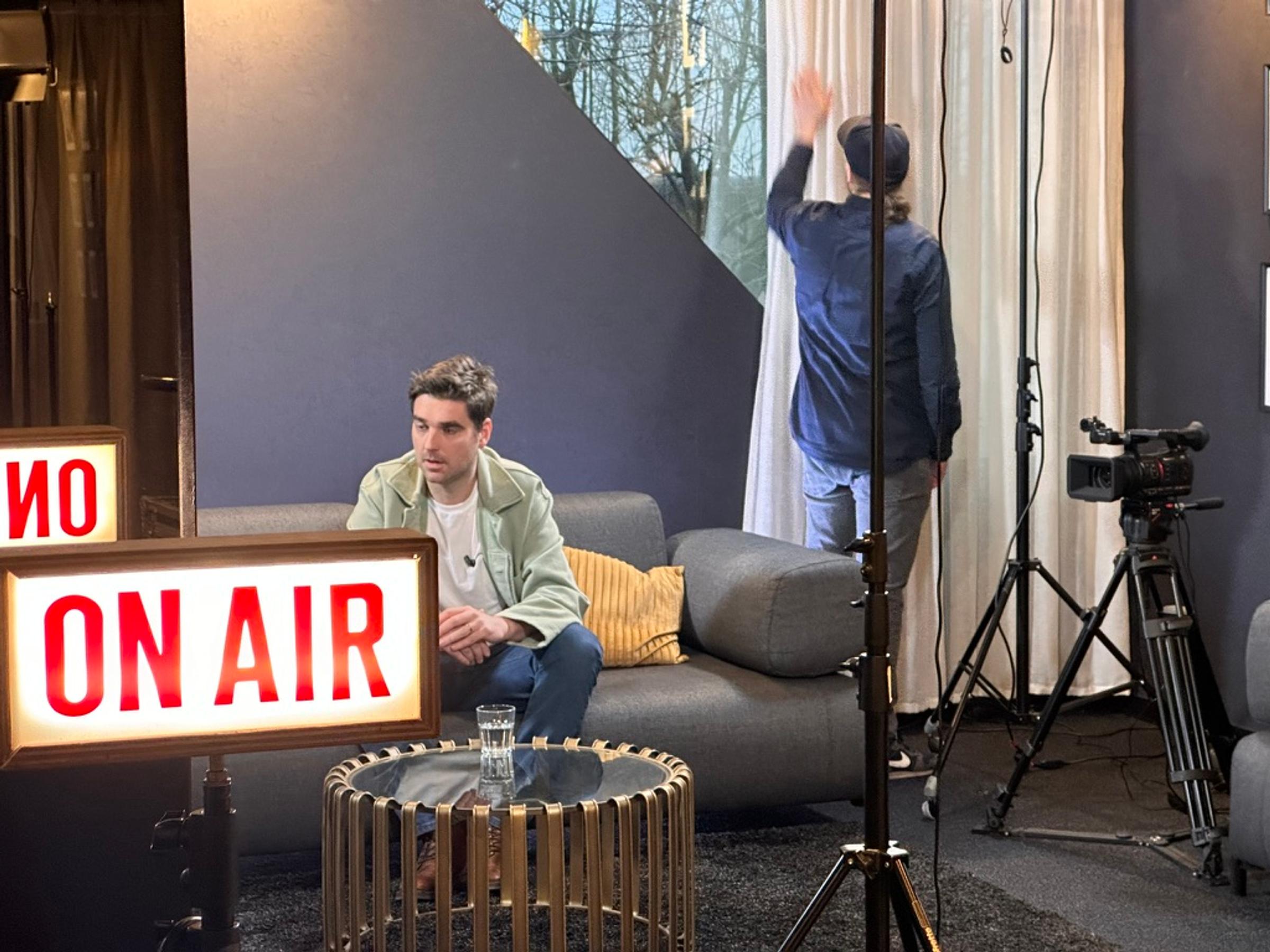 A podcast set with a lit ON AIR sign, a guest seated on the couch and a camera ready to roll