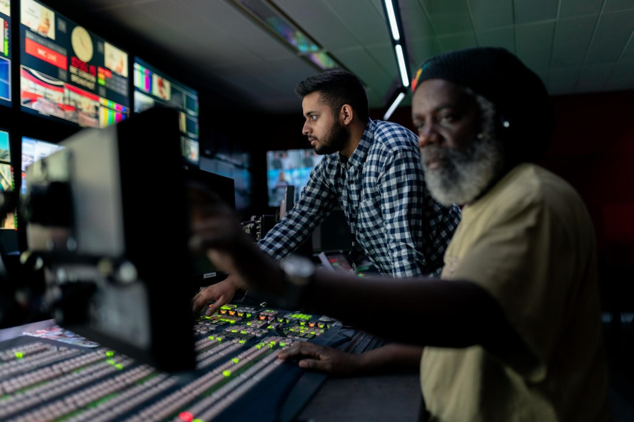 Two operators at a vision mixer in a broadcast control room, a wall of monitors showing live feeds behind them