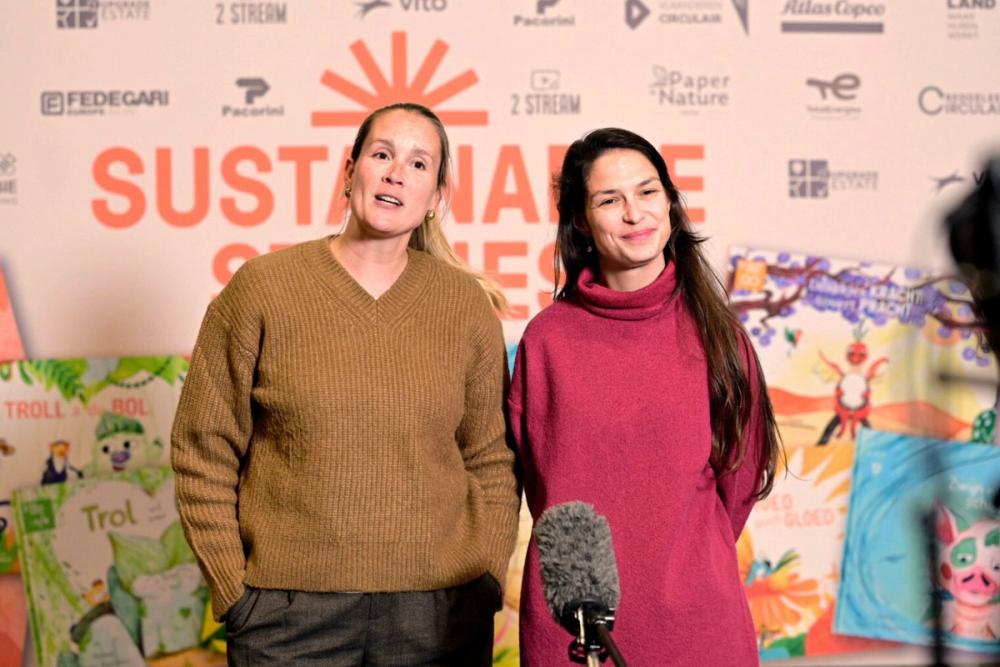 Two women interviewed at a Sustainable Stories event in front of a sponsor wall and children's book covers