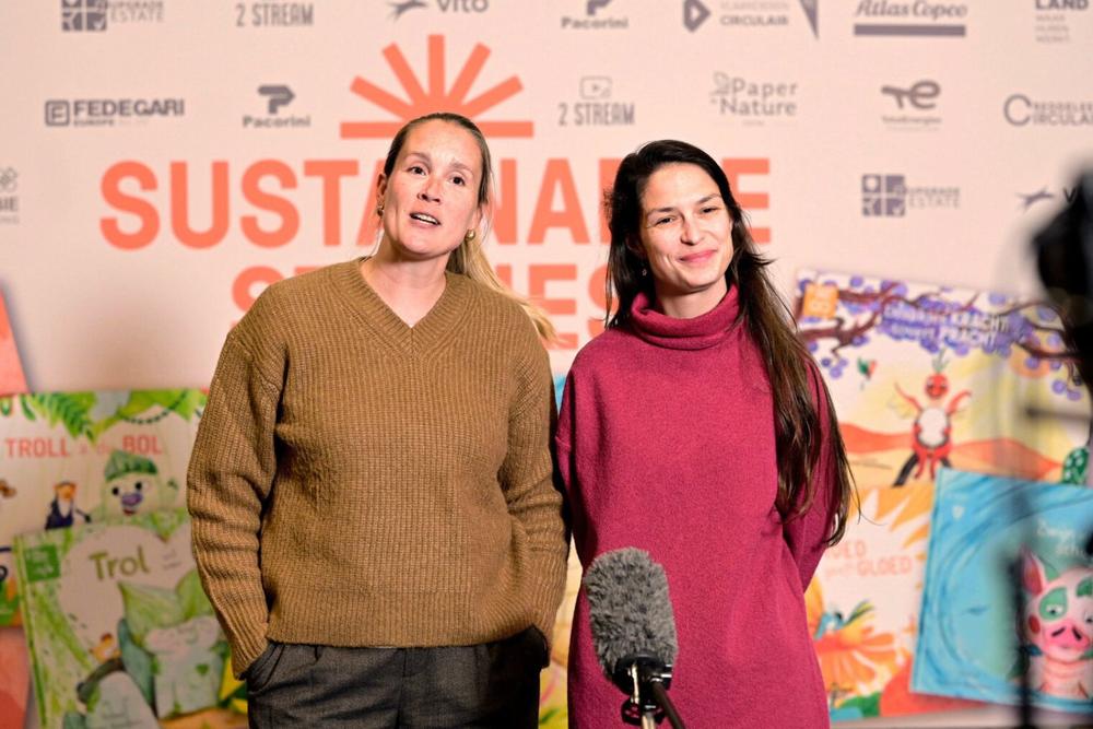 Two women interviewed at a Sustainable Stories event in front of a sponsor wall and children's book covers