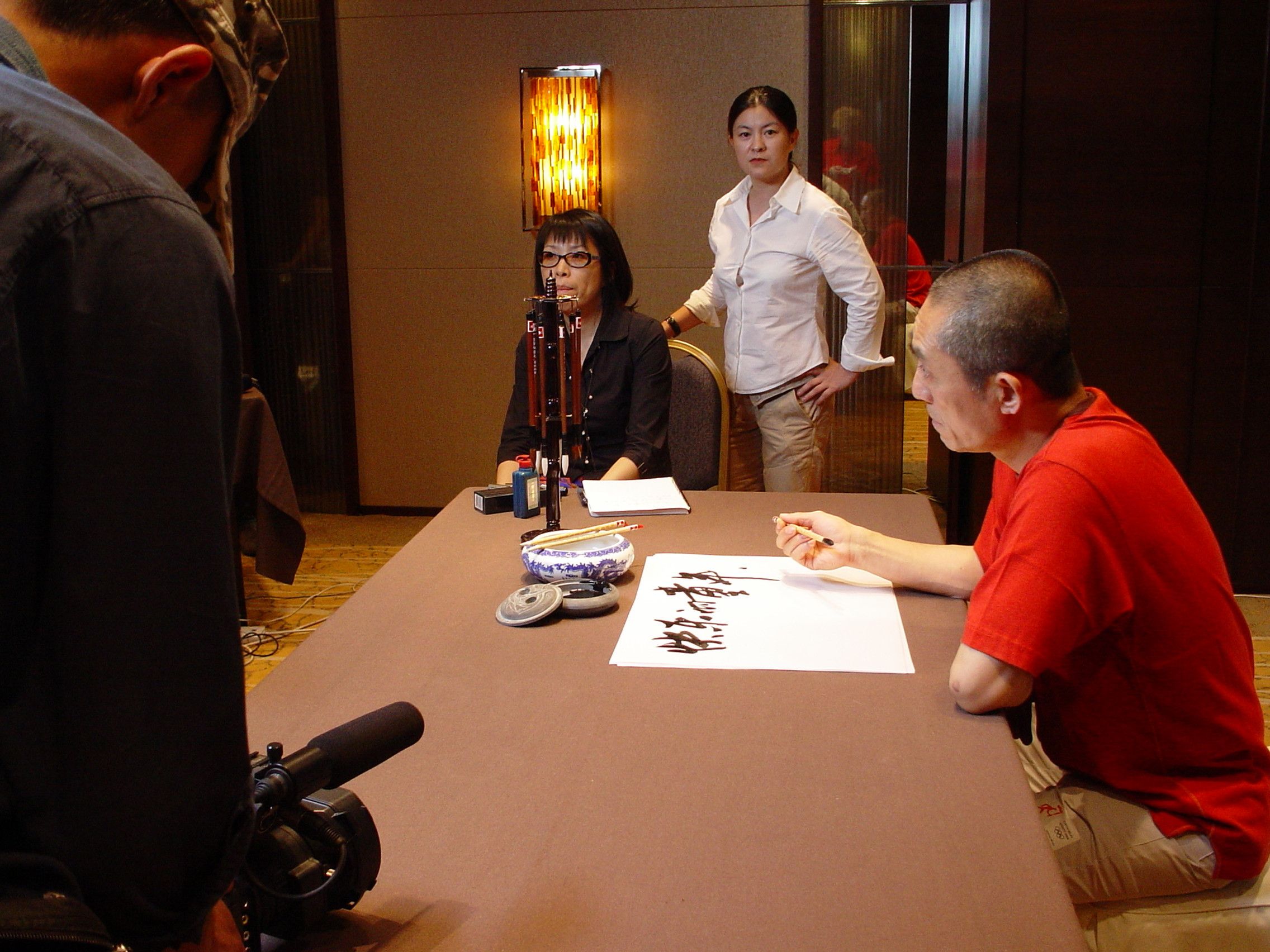 Man in a red T-shirt writing Chinese calligraphy at a table while a camera operator films in the foreground