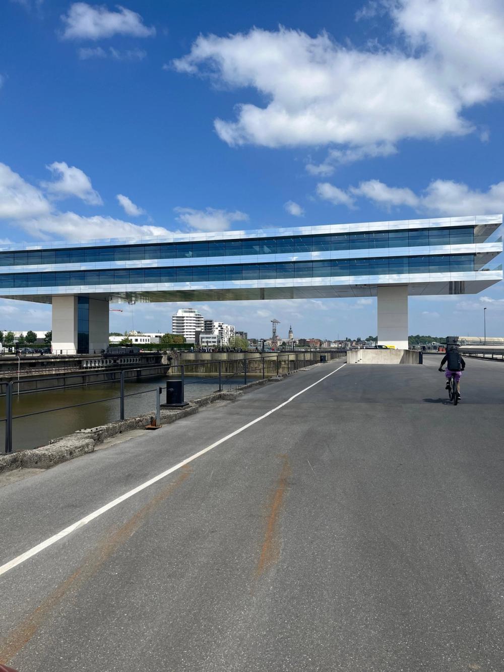 Cyclist on a quayside road beneath a long glass office building raised on two pillars