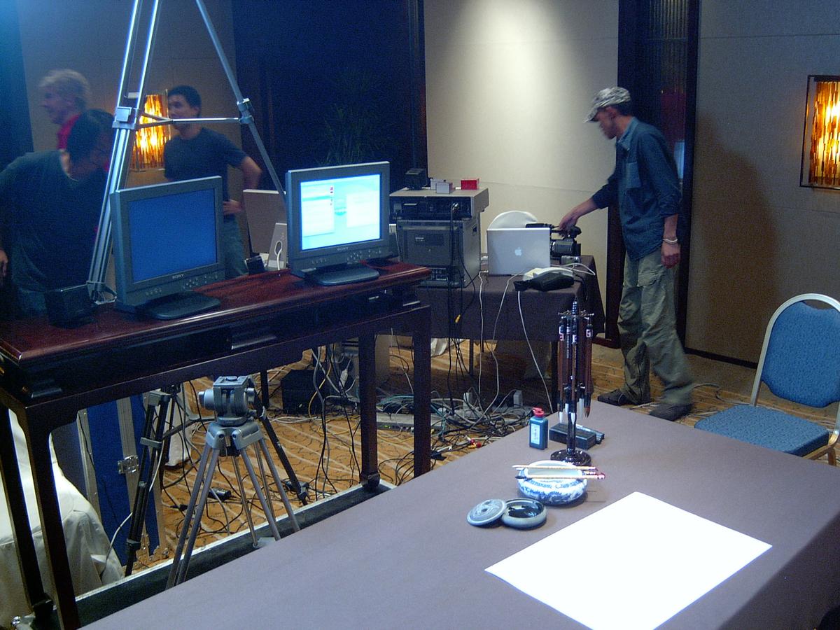 Streaming control table with two monitors, a MacBook and cabling beside a calligraphy table with brushes and ink