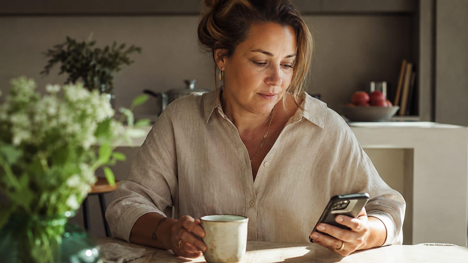 Frau Mitte 40 mit Kaffeetasse und Smartphone am Küchentisch – die digitale Sprechstunde des Menopause Zentrum lässt sich bequem und ortsunabhängig von zu Hause wahrnehmen.