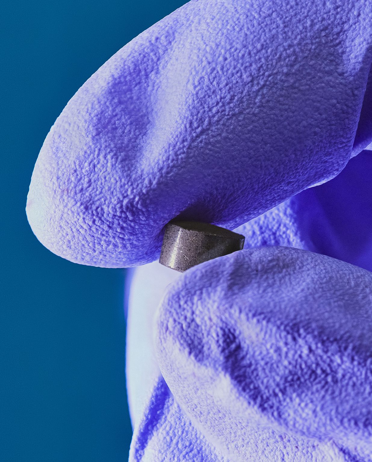 A hand in a purple glove holds a small catalyst pellet