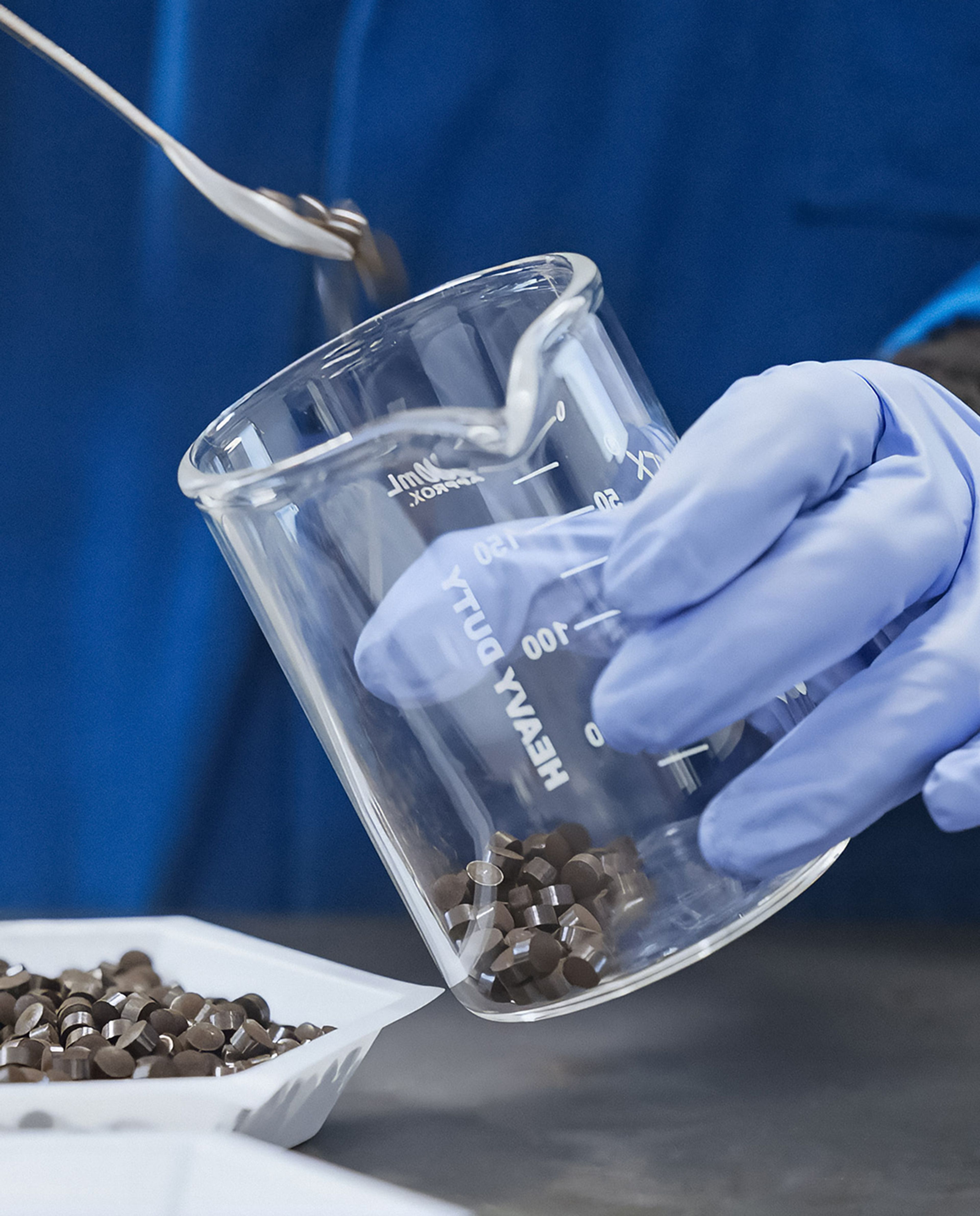A gloved hand spoons dark cylindrical pellets into a glass beaker.