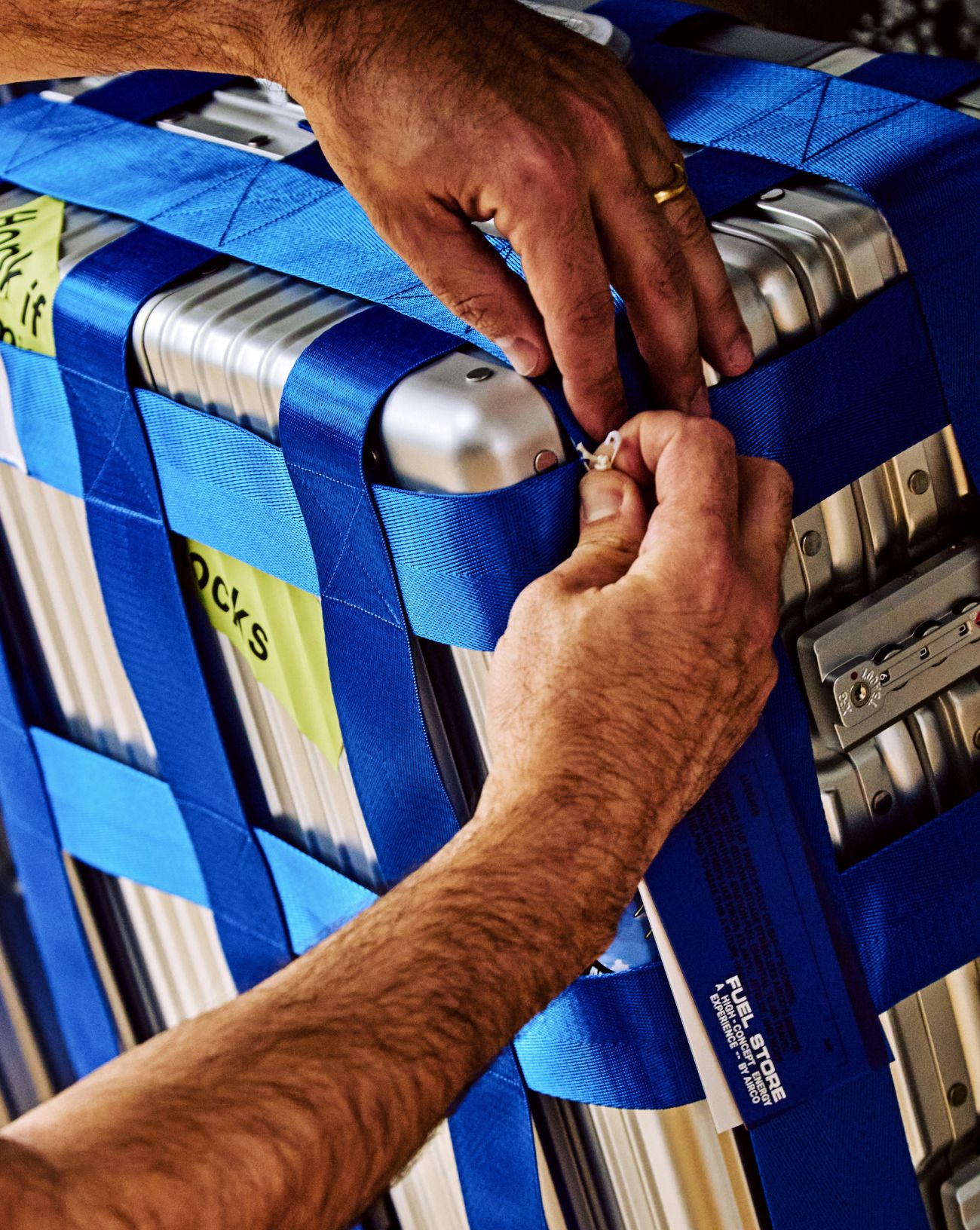 up-close shot of two hands putting a safety pin on blue straps that are on a silver suitcase