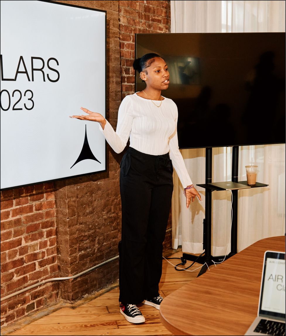 A woman of AIRCO's AIR Scholars Cohort 2 presents in front of a screen displaying a black triangular logo.
