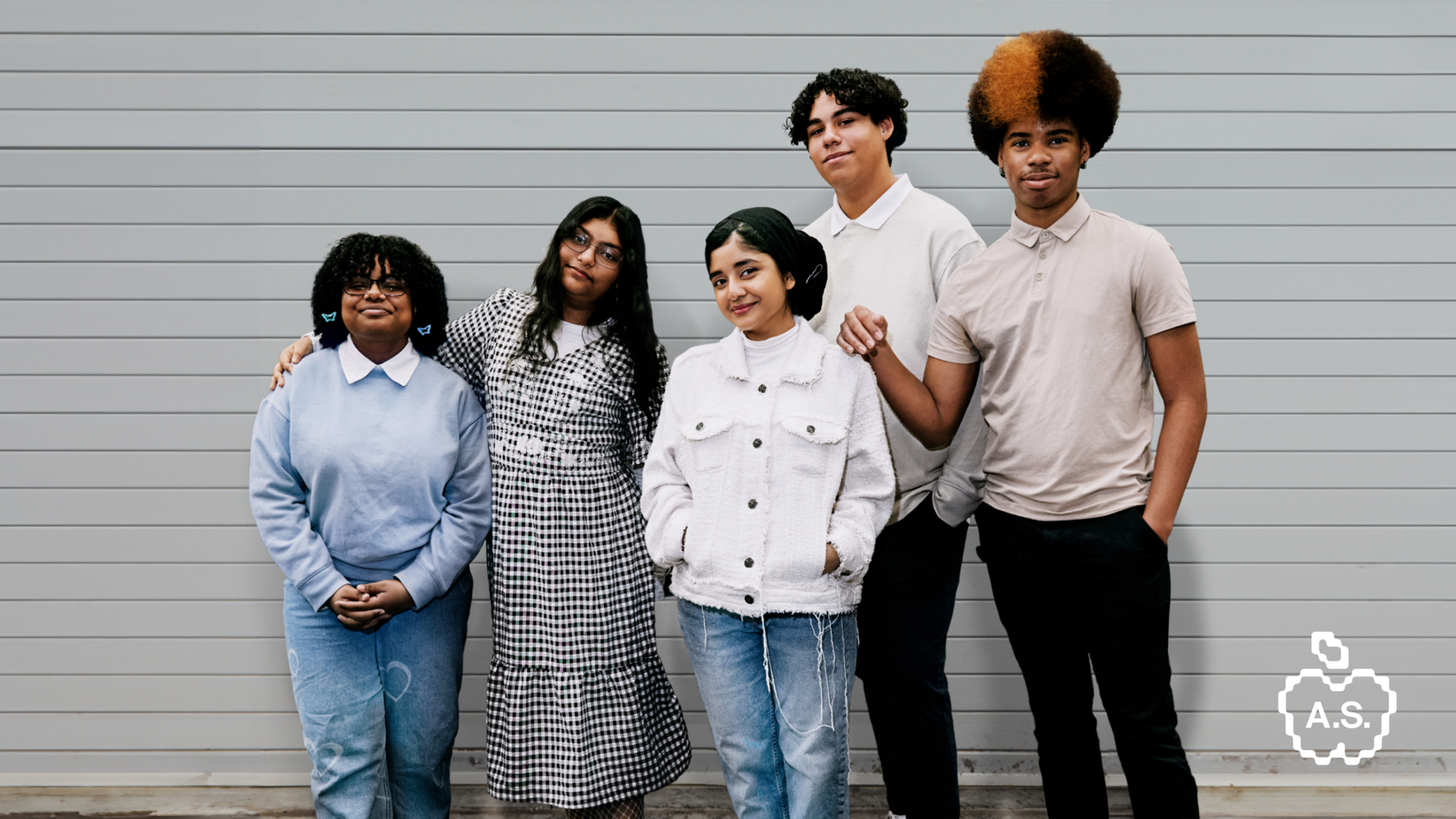 A diverse group of five young people in casual clothes stands smiling against a grey paneled wall; one person has a two-toned afro.