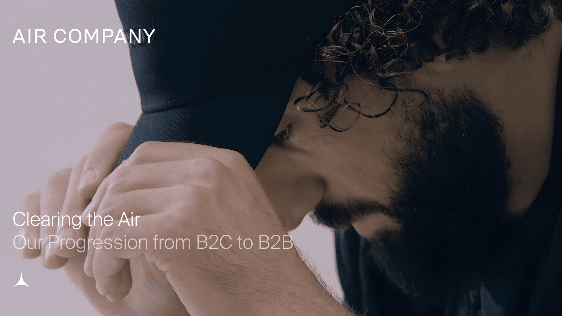 A person AIRCO (formerly Air Company) co-founder Gregory Constantine sitting in a chair. Image is zoomed in on his side profile and he is looking down while holding his hat. with curly hair and a beard looks down, hands holding a dark cap. Text reads: "AIR COMPANY Clearing the Air Our Progression from B2C to B2B."