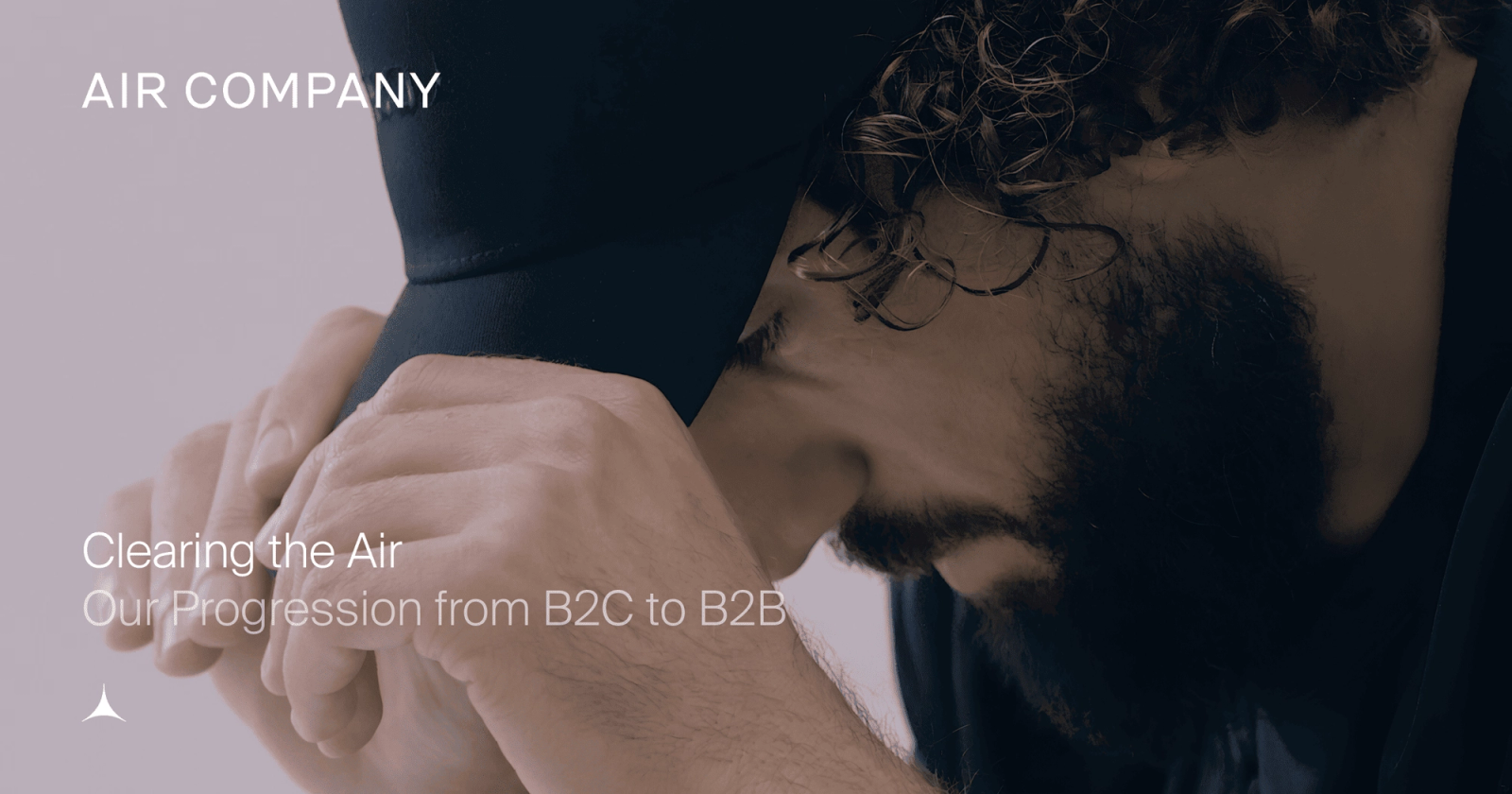 A person AIRCO (formerly Air Company) co-founder Gregory Constantine sitting in a chair. Image is zoomed in on his side profile and he is looking down while holding his hat. with curly hair and a beard looks down, hands holding a dark cap. Text reads: "AIR COMPANY Clearing the Air Our Progression from B2C to B2B."