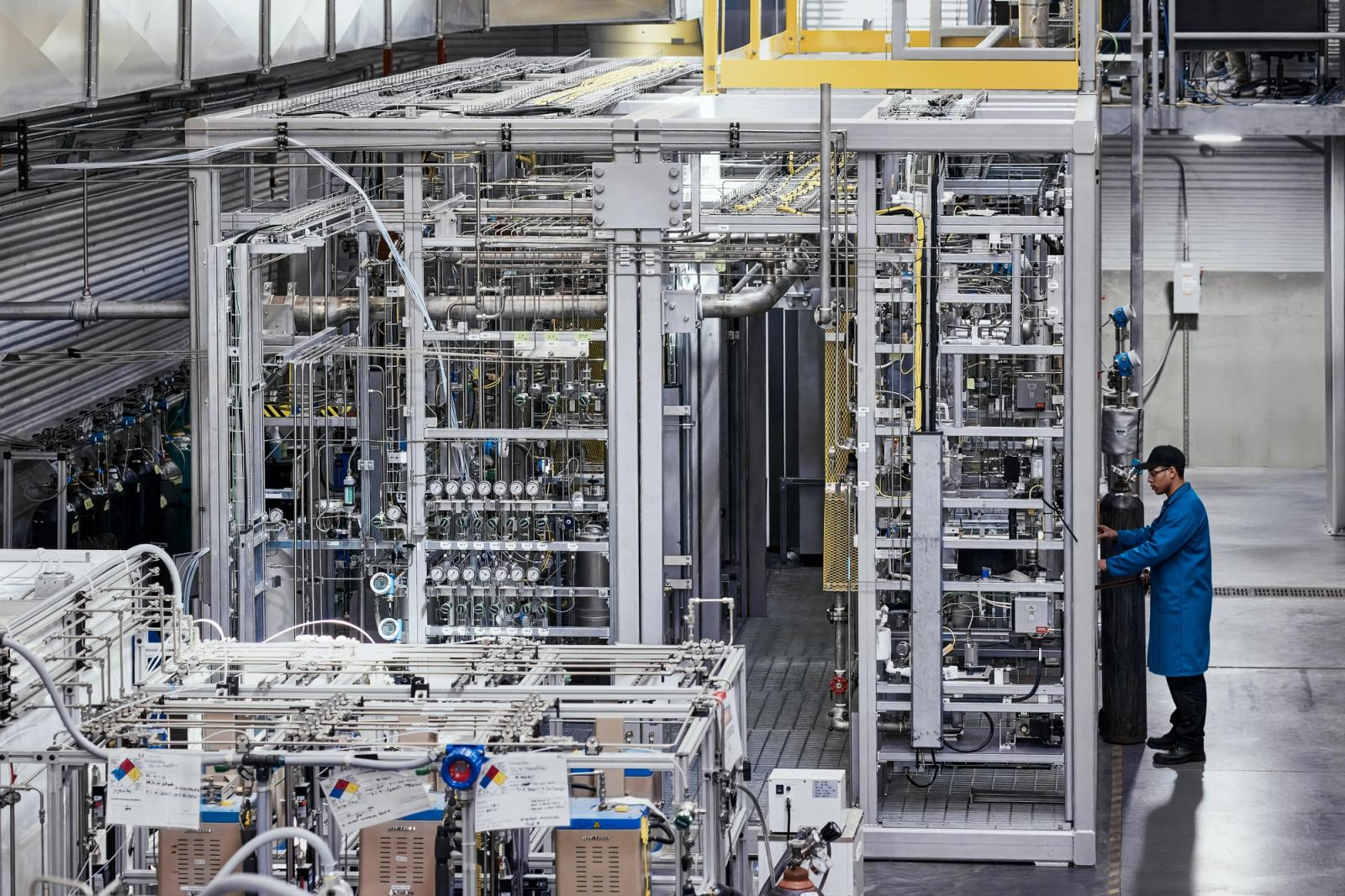 A person in a blue lab coat handles a gas cylinder amidst complex industrial machinery.
