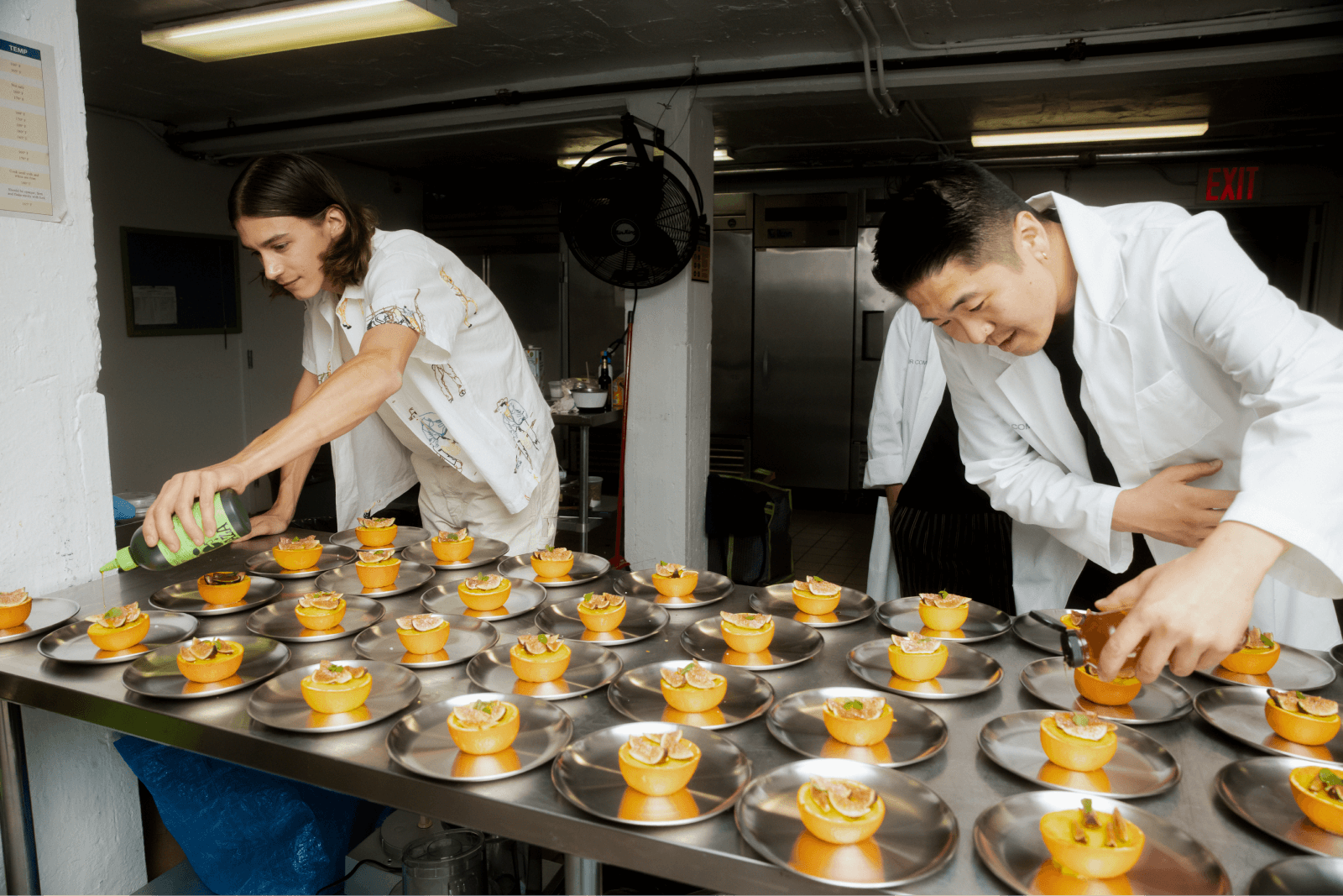 two men in a stainless steel kitchen preparing an orange dessert 