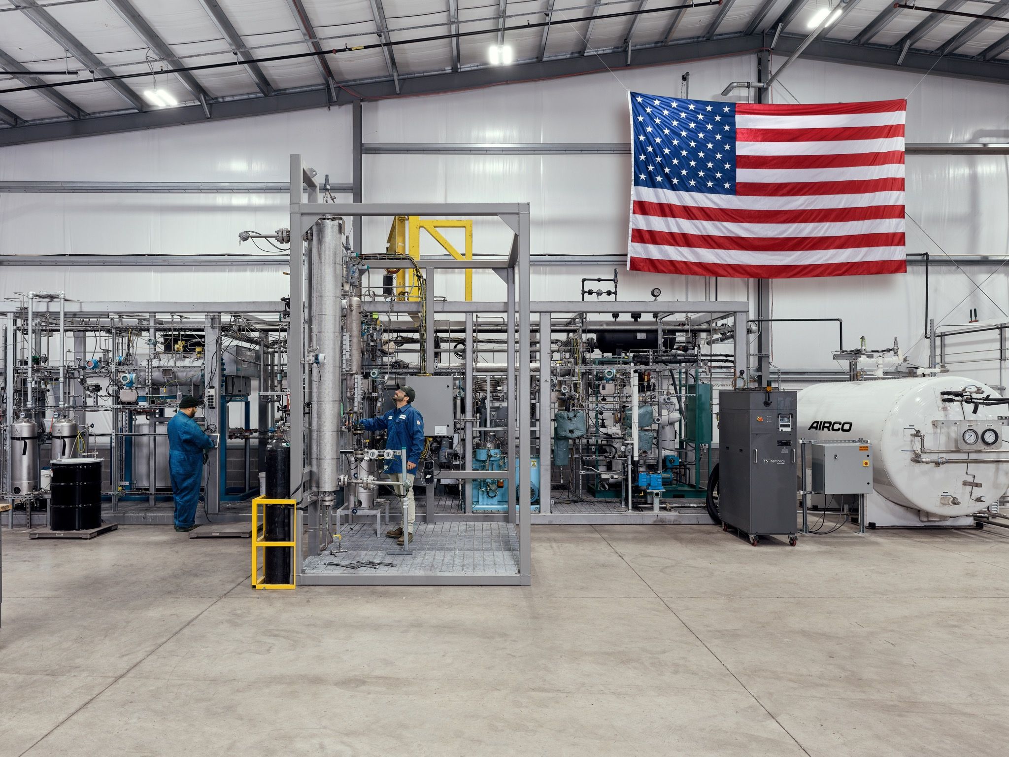 two men in blue suits working in a tech facility with an American flag behind them