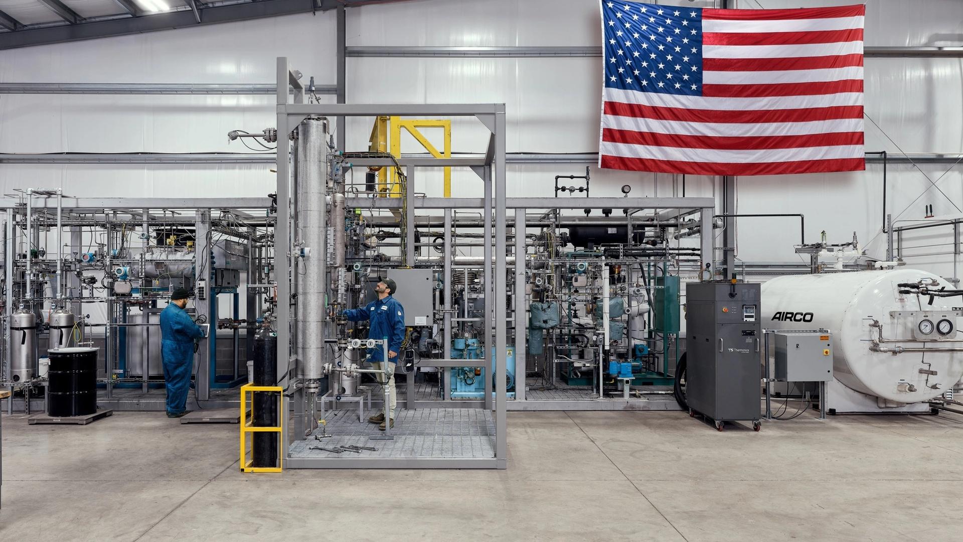two men in blue suits working in a tech facility with an American flag behind them