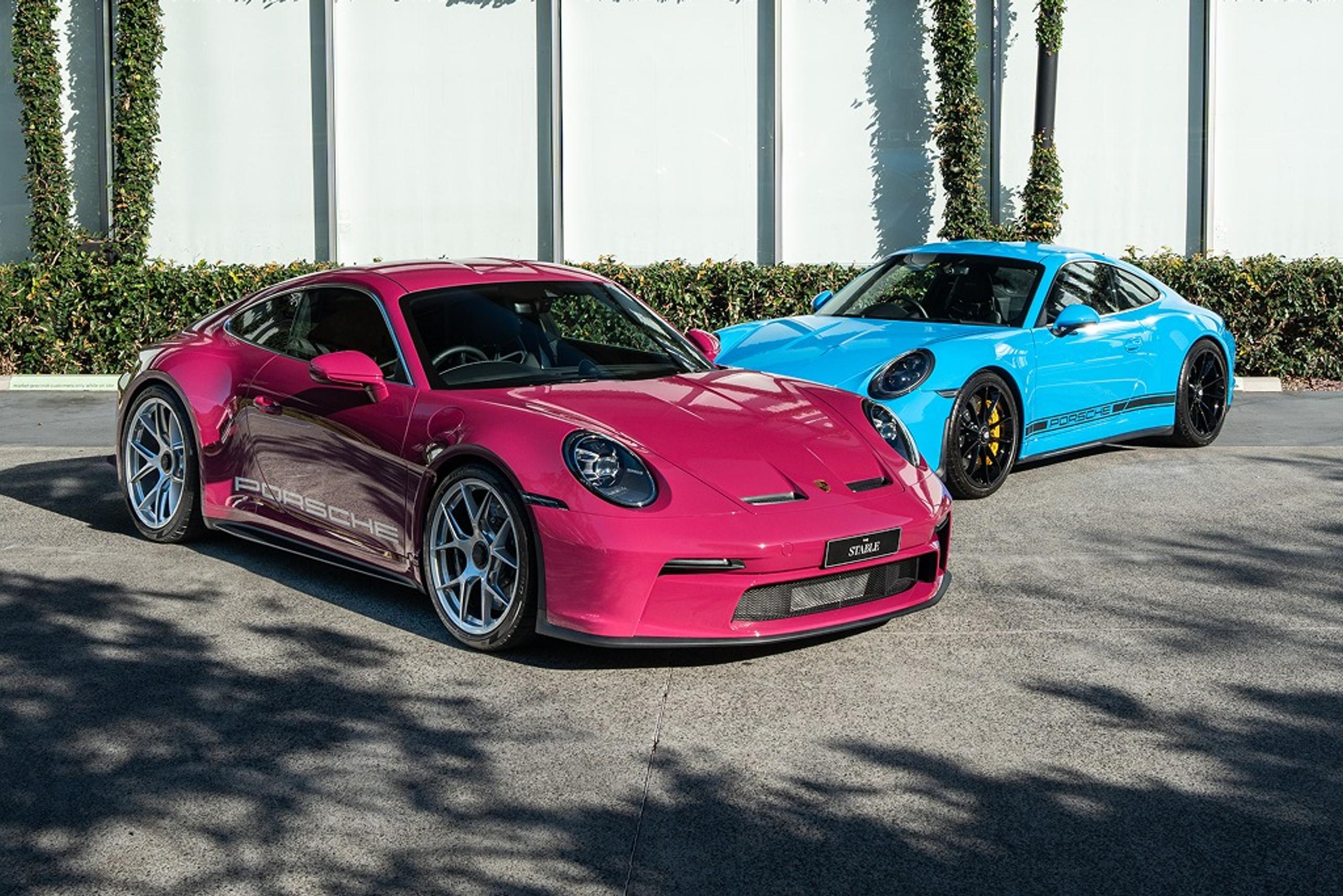 A pink Porsche 911 and a blue Porsche 911 parked in open shade in front of a white wall covered in a green climbing-plant trellis
