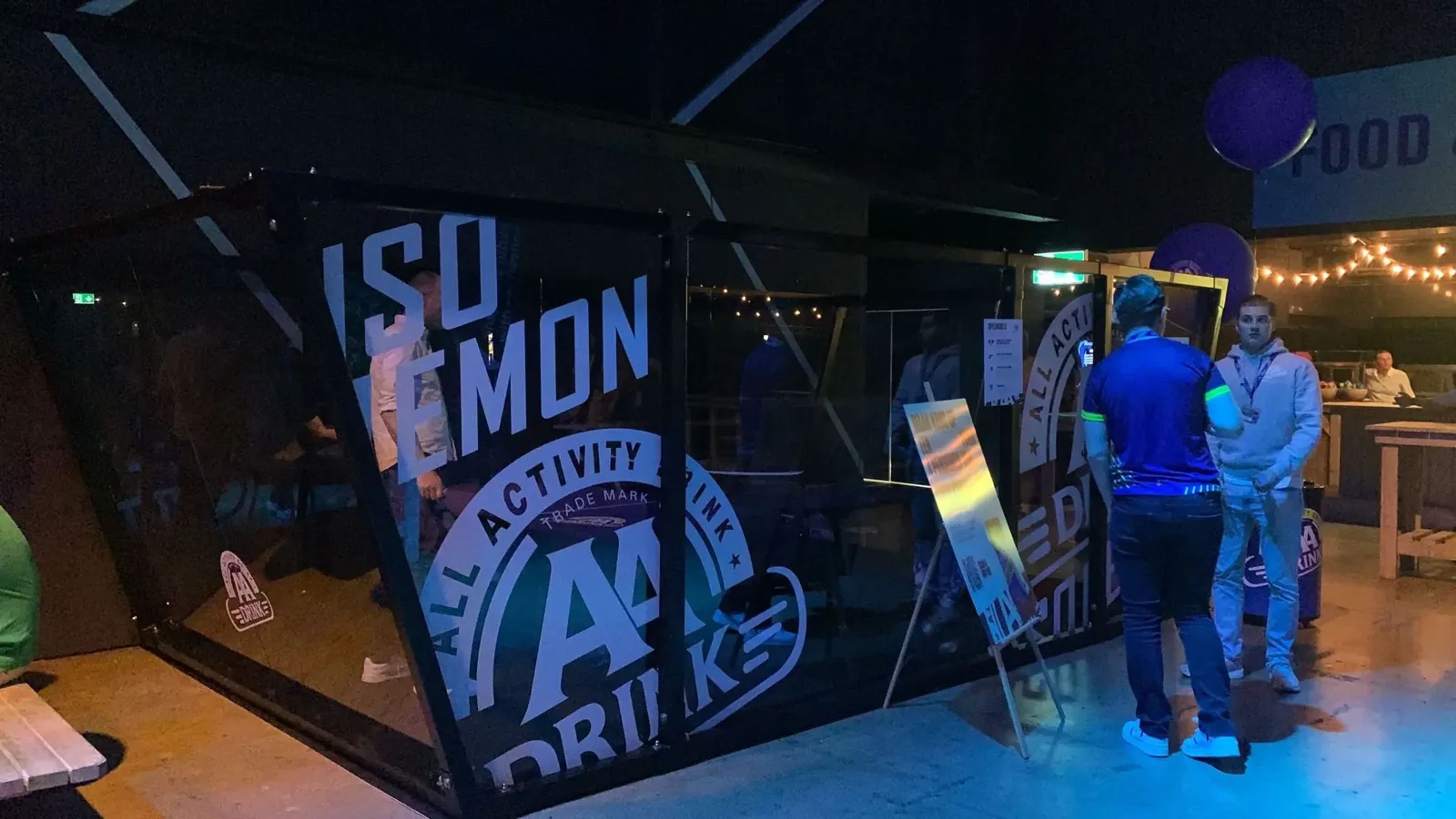 Glazen kooi van de Padel Ping Pong-baan met AA Drink-opdruk, met bezoekers op een evenement