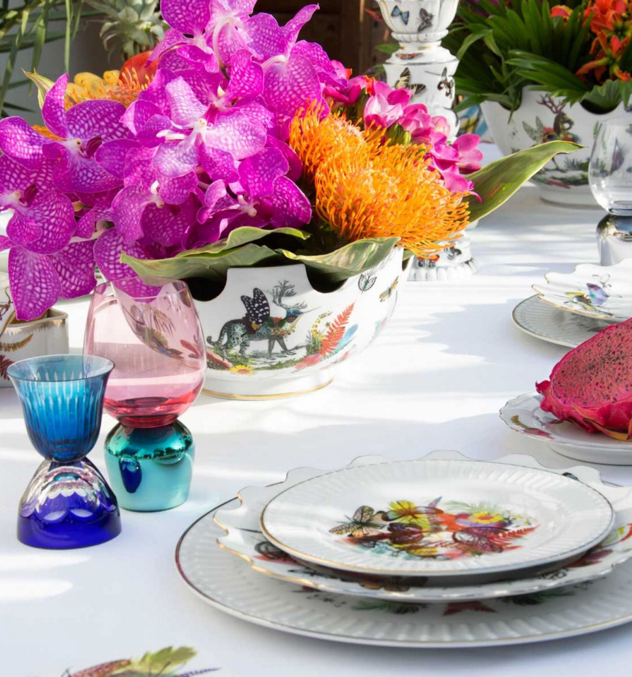Orchideás virágkompozíció, mintás porcelán tányérok és színes poharak terített asztalon